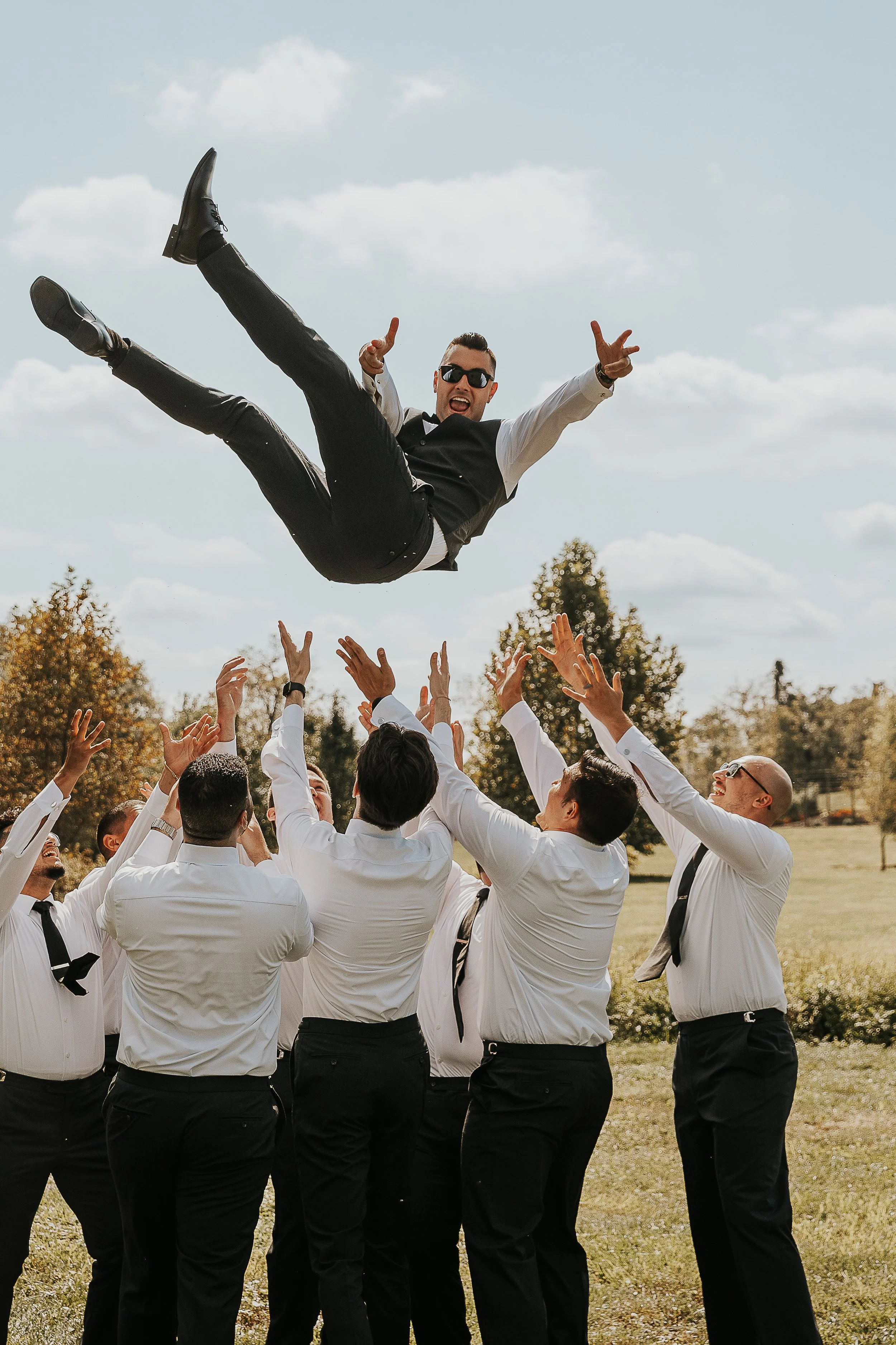 A man in sunglasses and a vest is being tossed into the air by a group of people dressed in white shirts and black pants, outdoors on a sunny day with trees in the background.