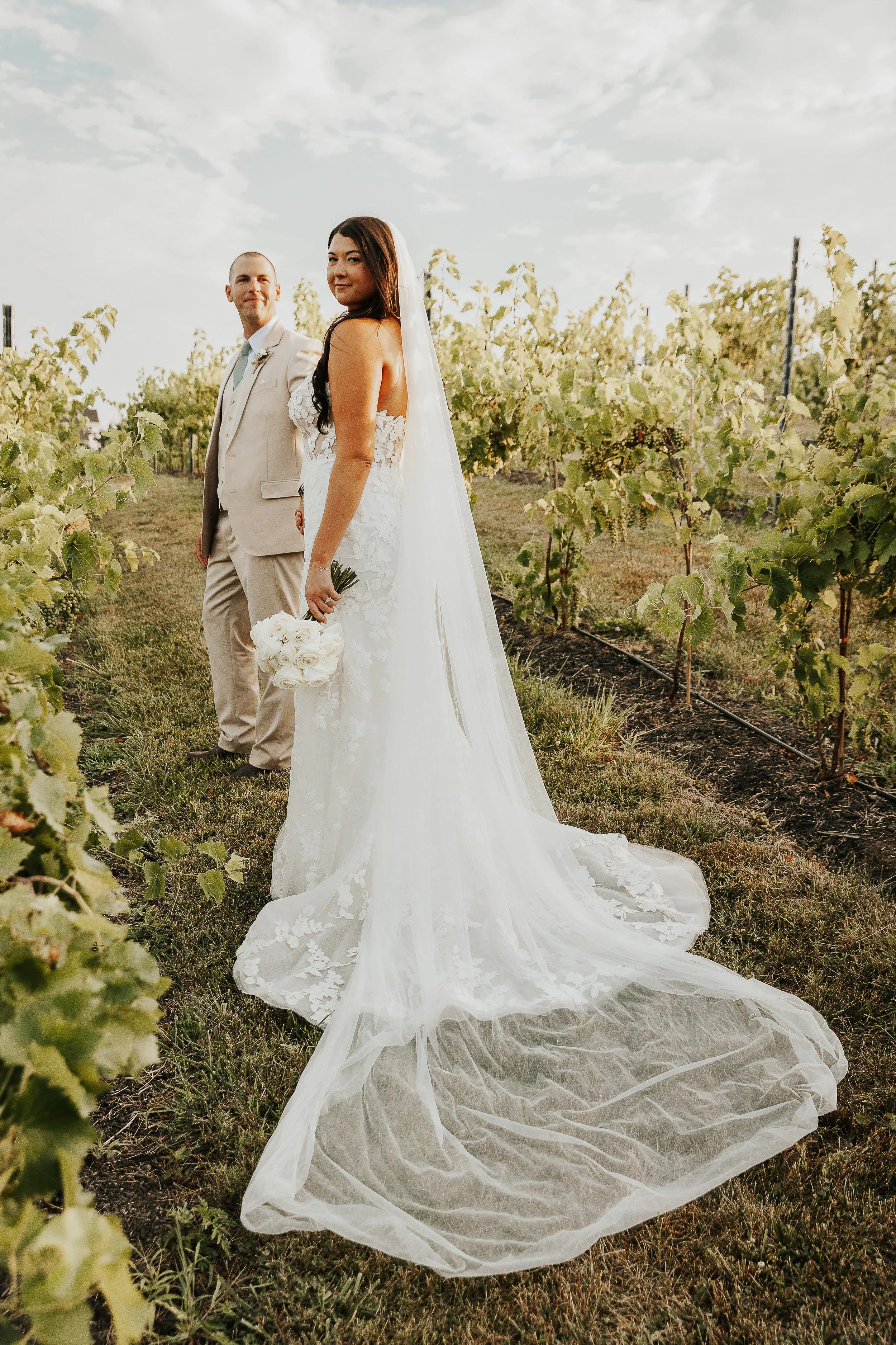 A bride and groom standing in a vineyard during sunset, with the bride holding a bouquet of white roses, both dressed in wedding attire, facing away from the camera but looking toward each other.