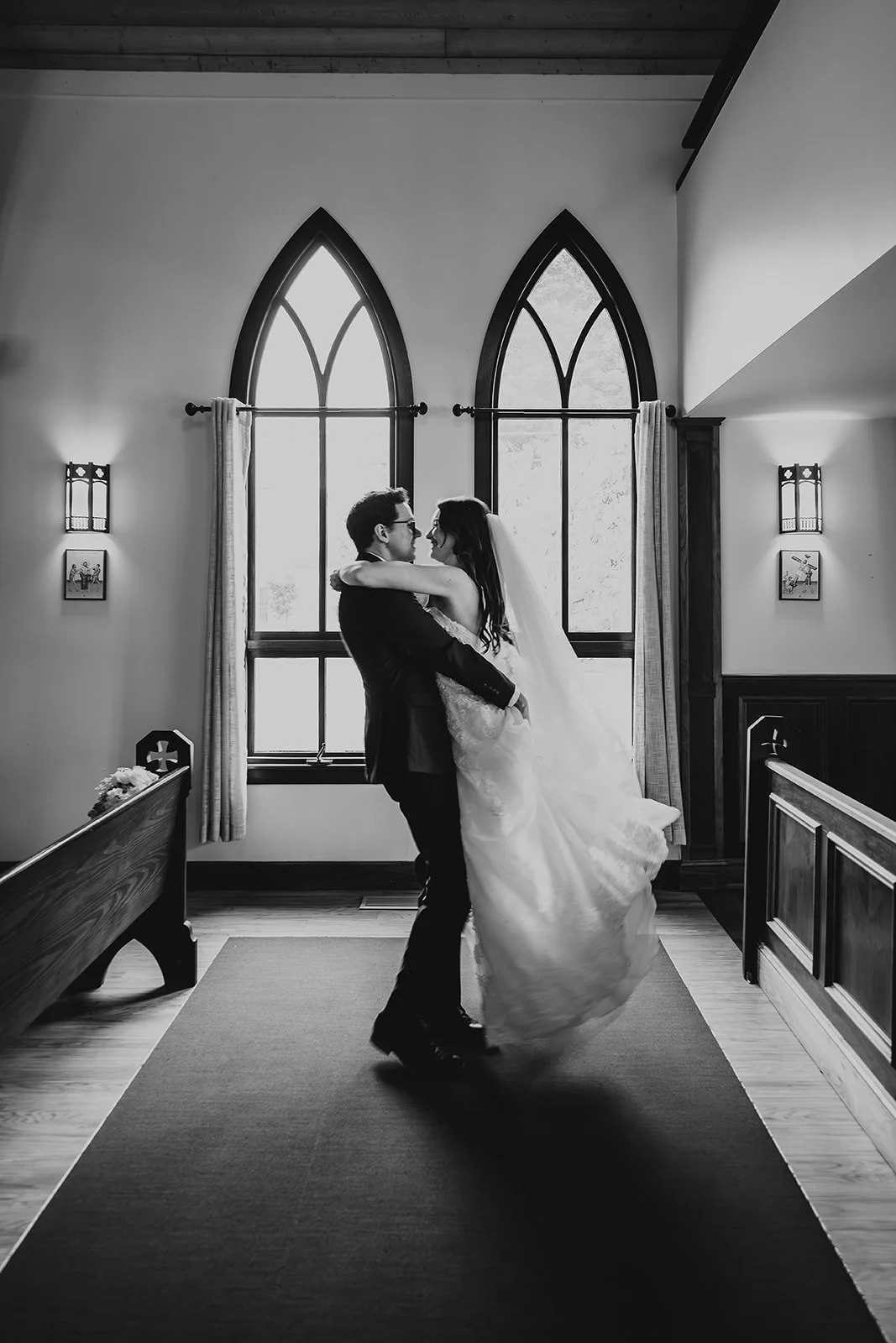 A black-and-white photo of a bride and groom dancing inside a church with large arched windows.