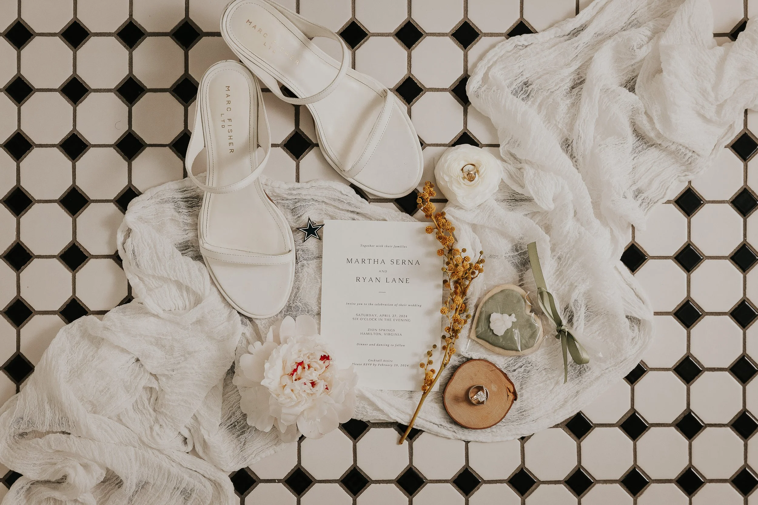 White heeled sandals, a wedding invitation, flowers, a ring, and decorative items arranged on a white textured cloth on a black and white tiled floor.