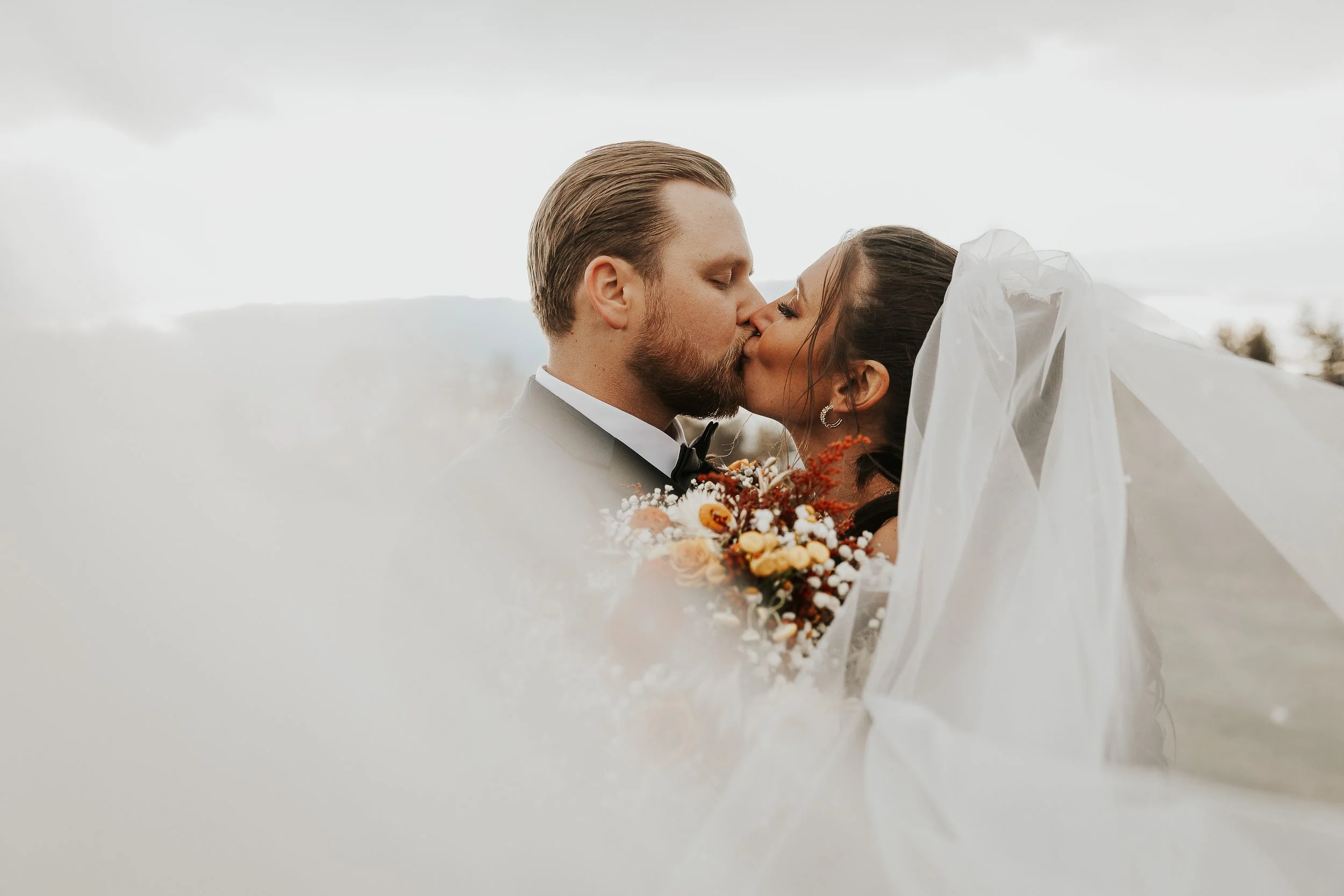 A bride and groom kissing outdoors, with the bride holding a bouquet of flowers.