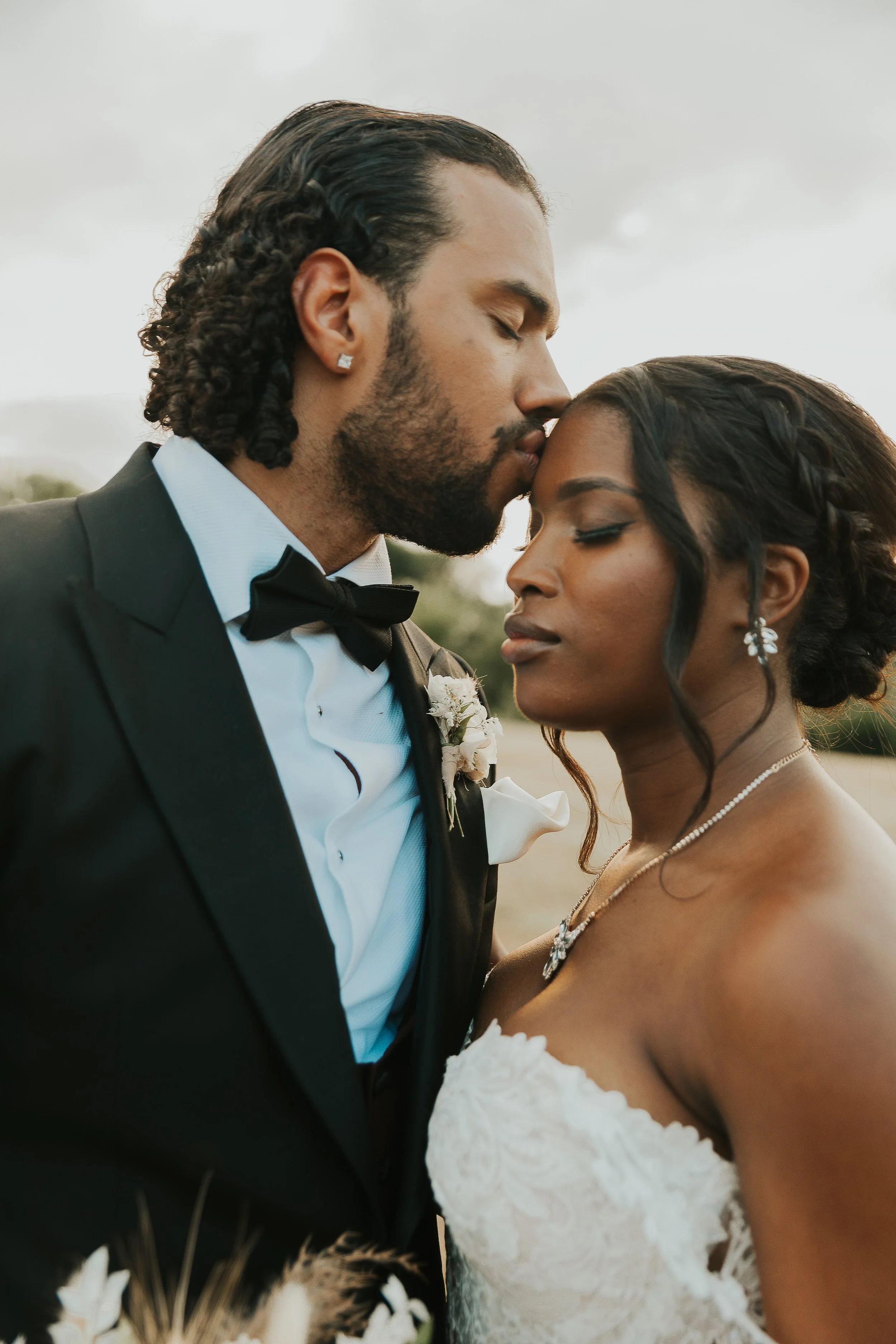 A man and a woman share a tender moment with foreheads touching, dressed in formal wedding attire, outside during daylight.