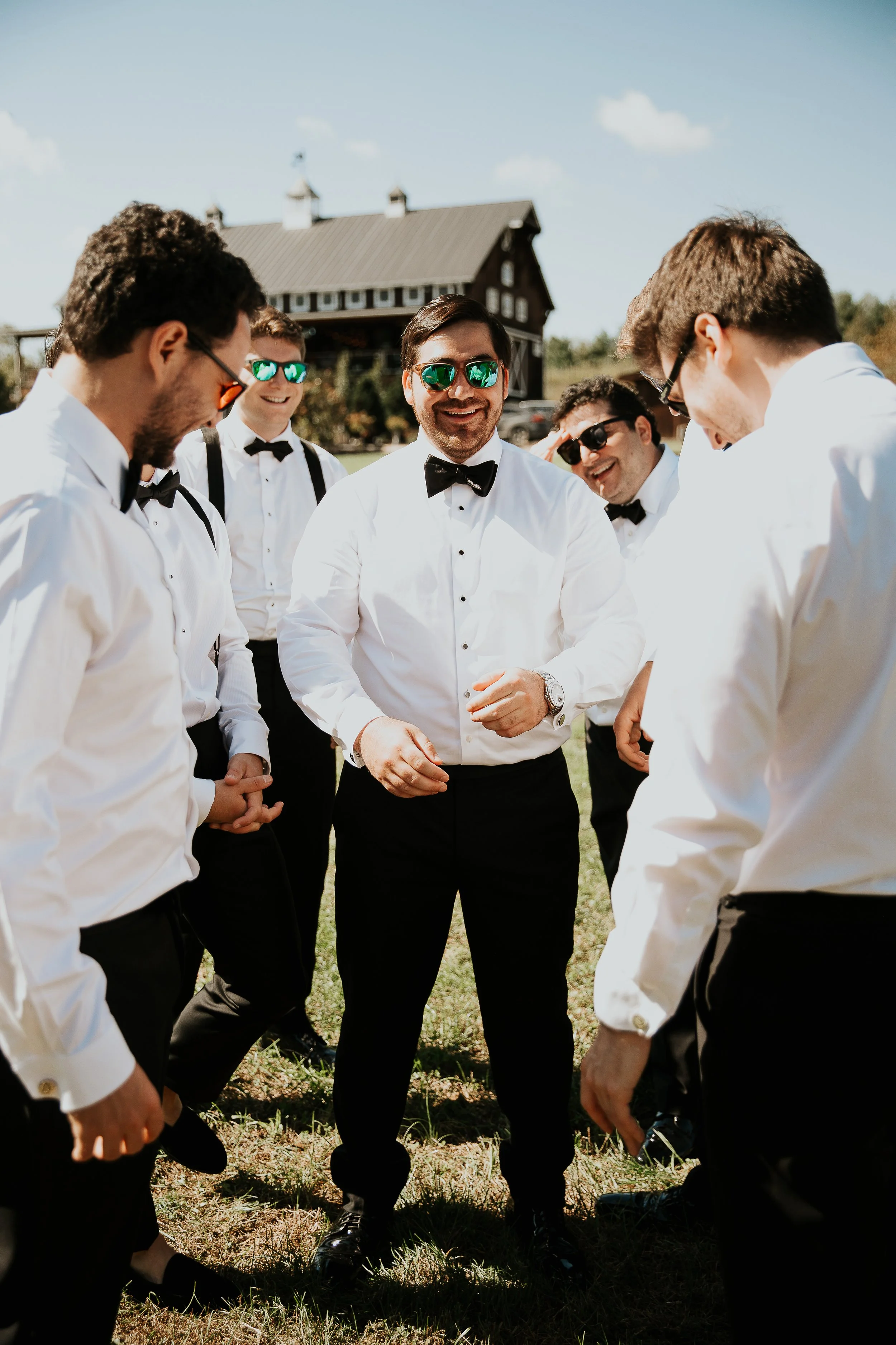 A group of six men dressed in formal white shirts, black bow ties, and suspenders, standing outdoors on a sunny day, likely at a wedding or formal event, with a rustic barn in the background.