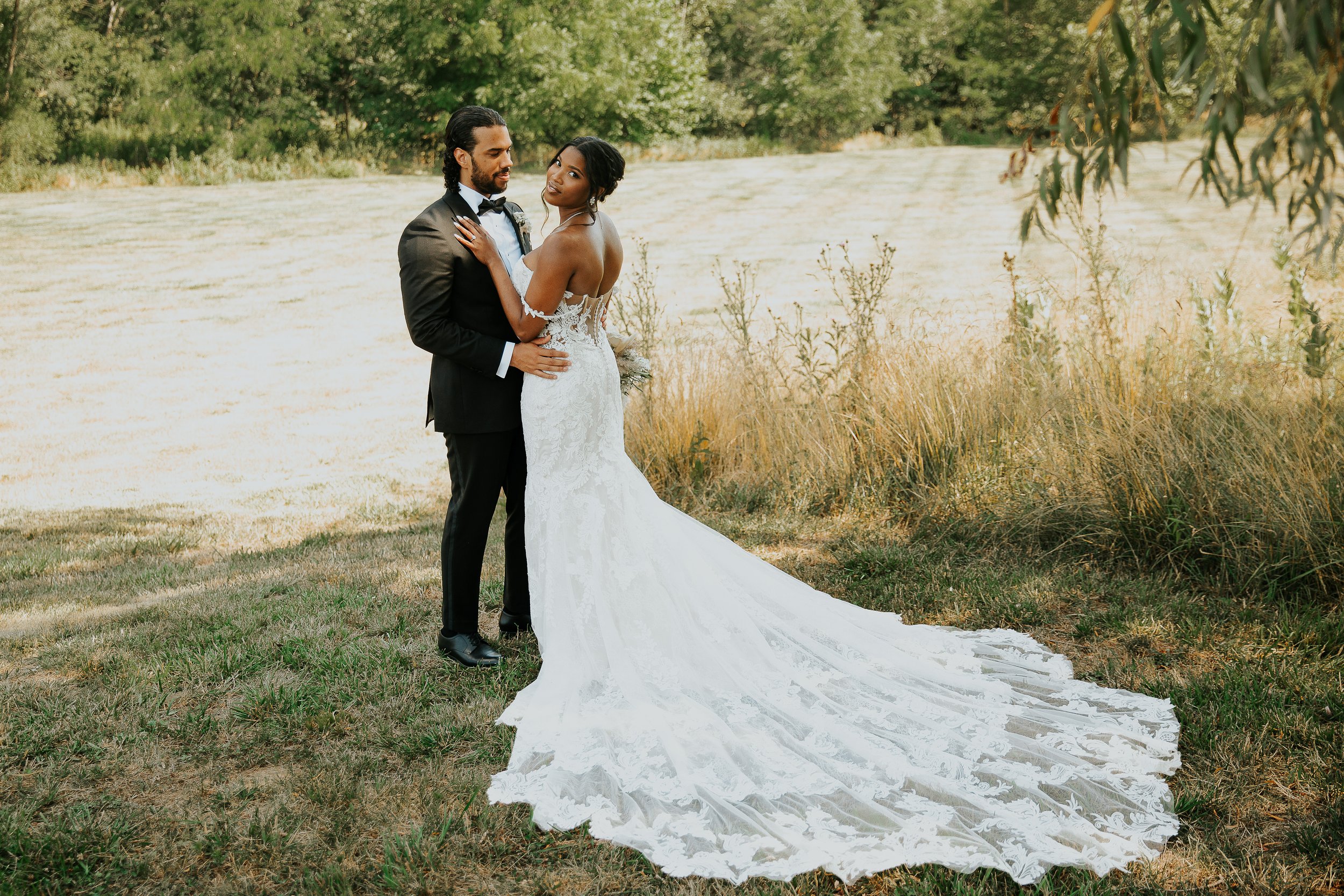 A bride and groom standing outdoors in a grassy area beside a body of water with trees in the background, the bride in a white wedding dress and the groom in a black tuxedo, posing for a wedding photo.