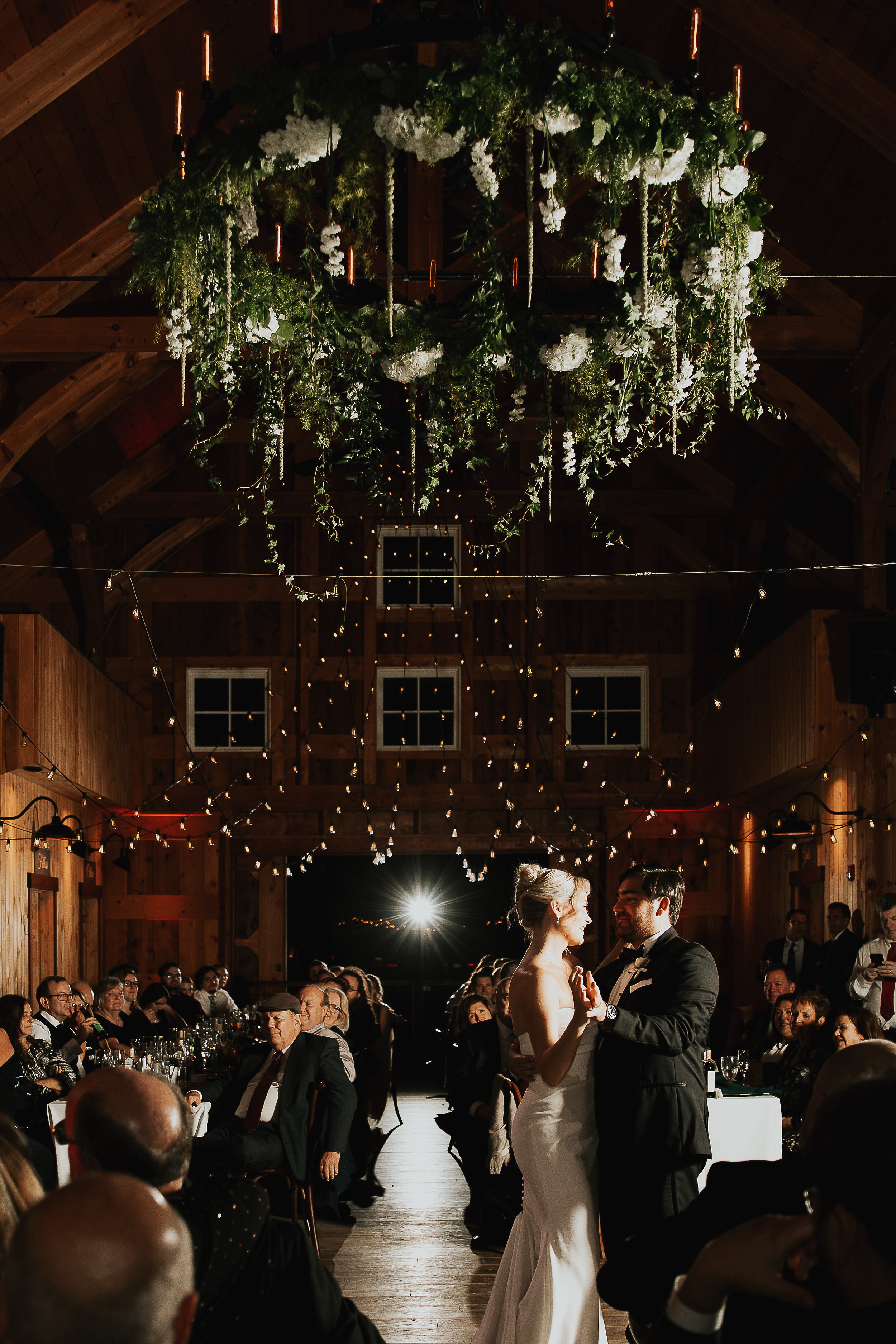 A bride and groom dancing during their wedding reception in a rustic wooden barn decorated with greenery and white flowers hanging from the ceiling, with guests seated around tables watching them.