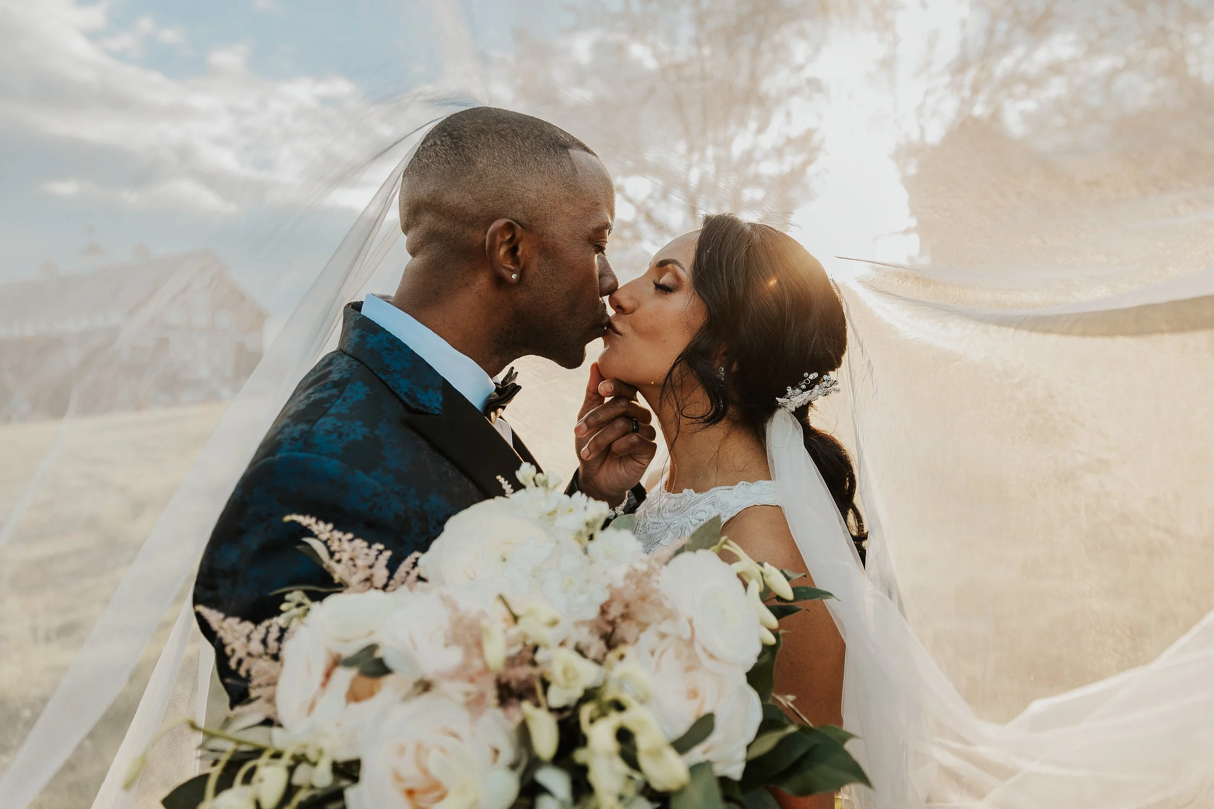 A bride and groom share a kiss under a wedding veil, with a bouquet of white flowers, outdoors during the day.