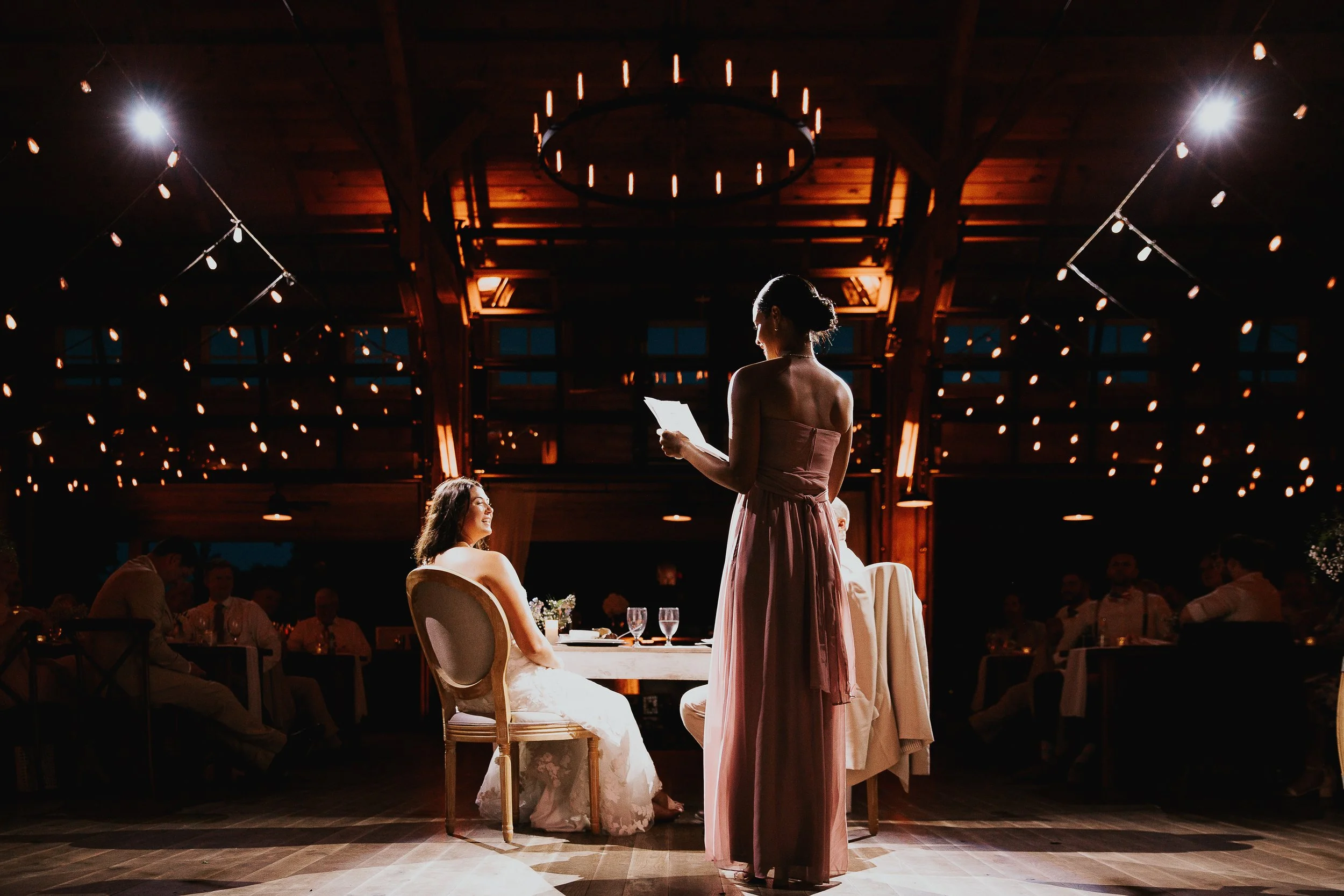A woman in a strapless pink dress reads from a paper while standing at a wedding reception, with the bride seated at a table and guests sitting around in a warmly lit room with string lights and a wooden ceiling.