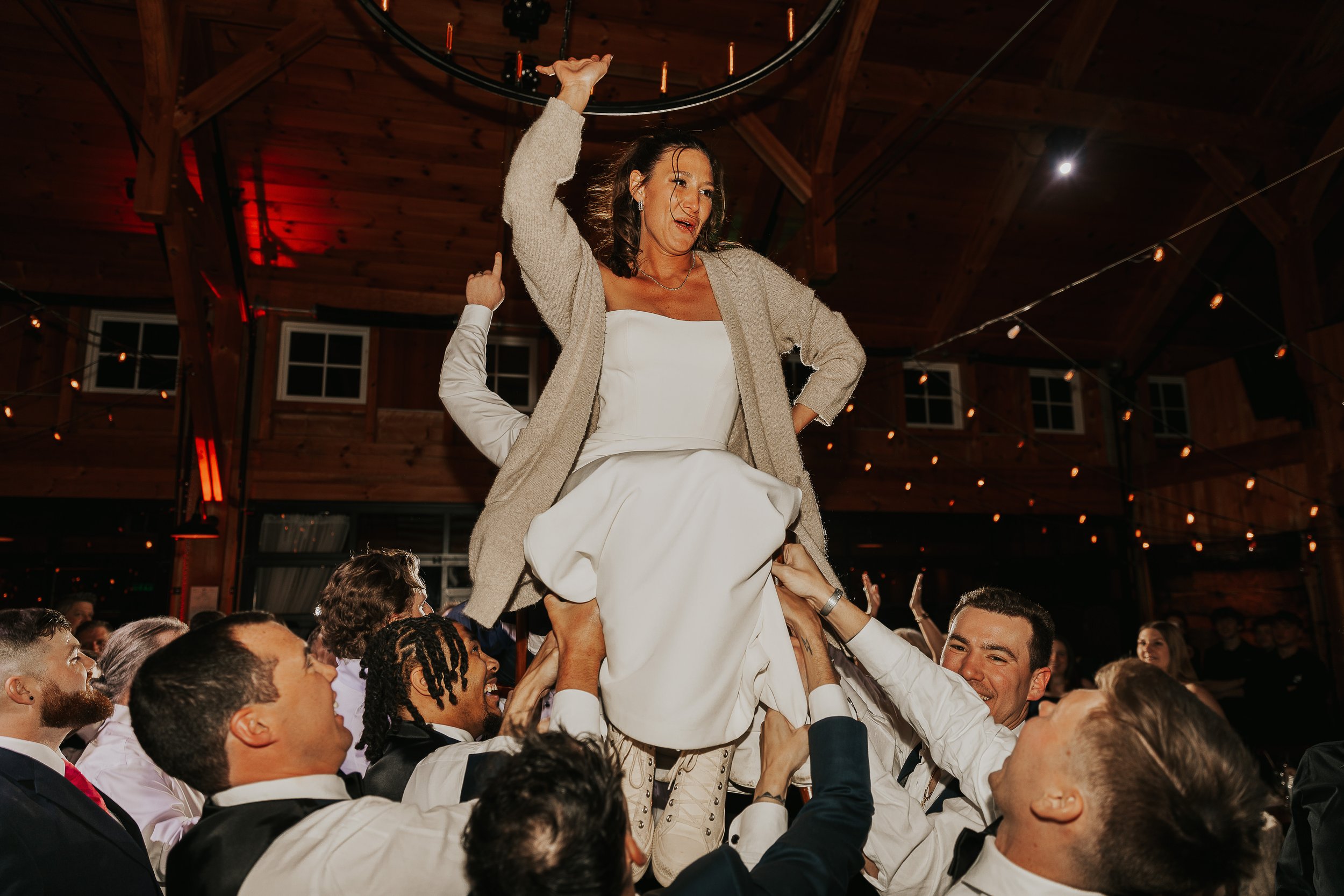 A woman in a white dress and beige cardigan is being lifted by a group of people at a celebration or wedding reception inside a wooden venue with string lights and windows in the background.