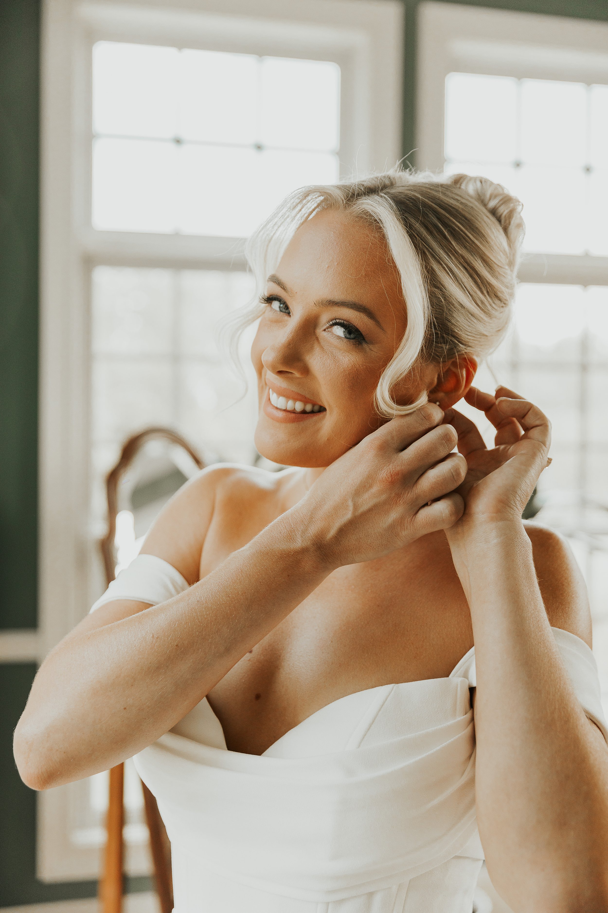 A woman with blonde hair styled in an updo, smiling and adjusting an earring, wearing a white off-shoulder dress, in a room with large windows in the background.