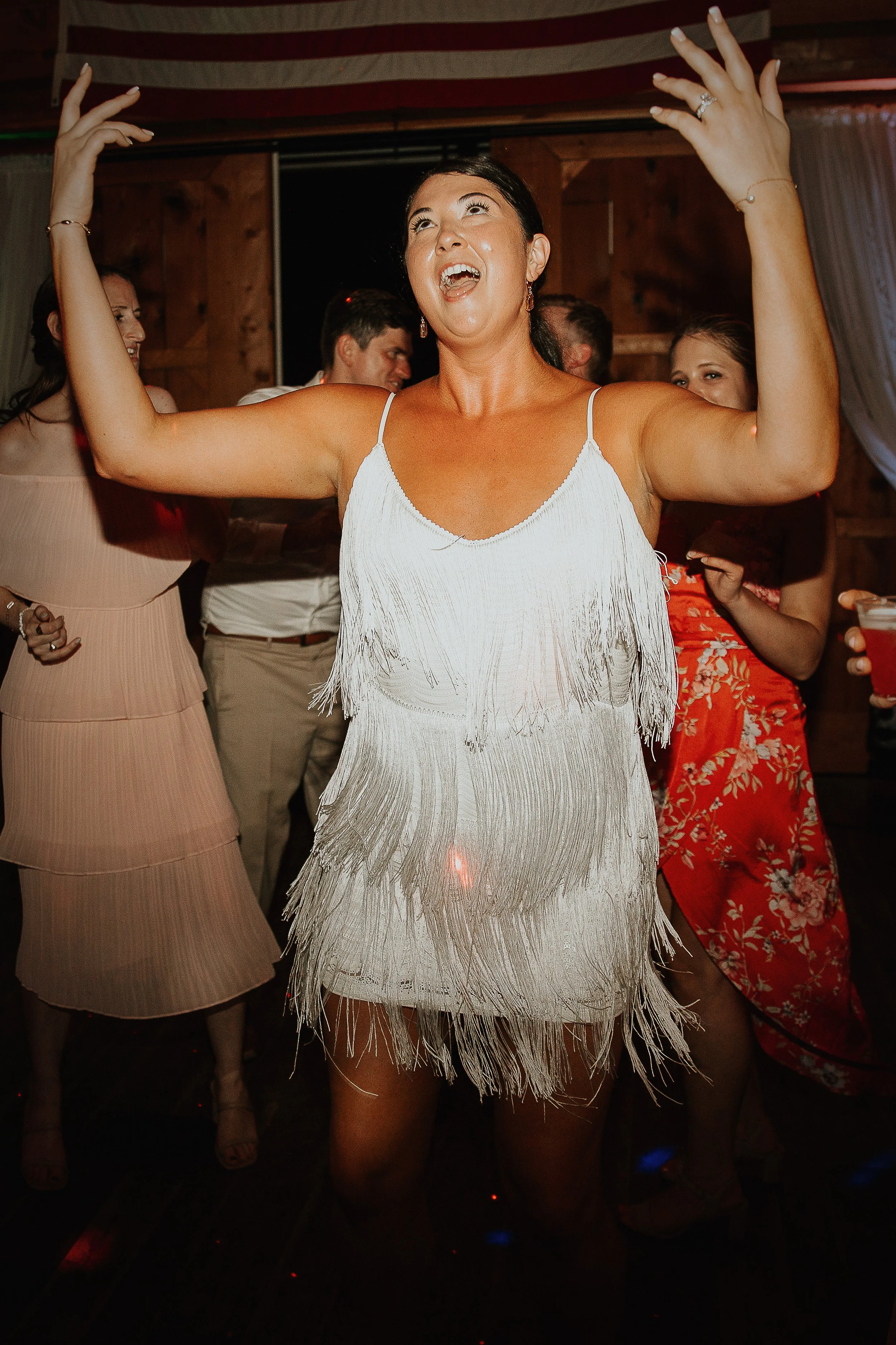 A woman in a white fringed dress dancing and smiling at a celebration or party, with other people in the background.