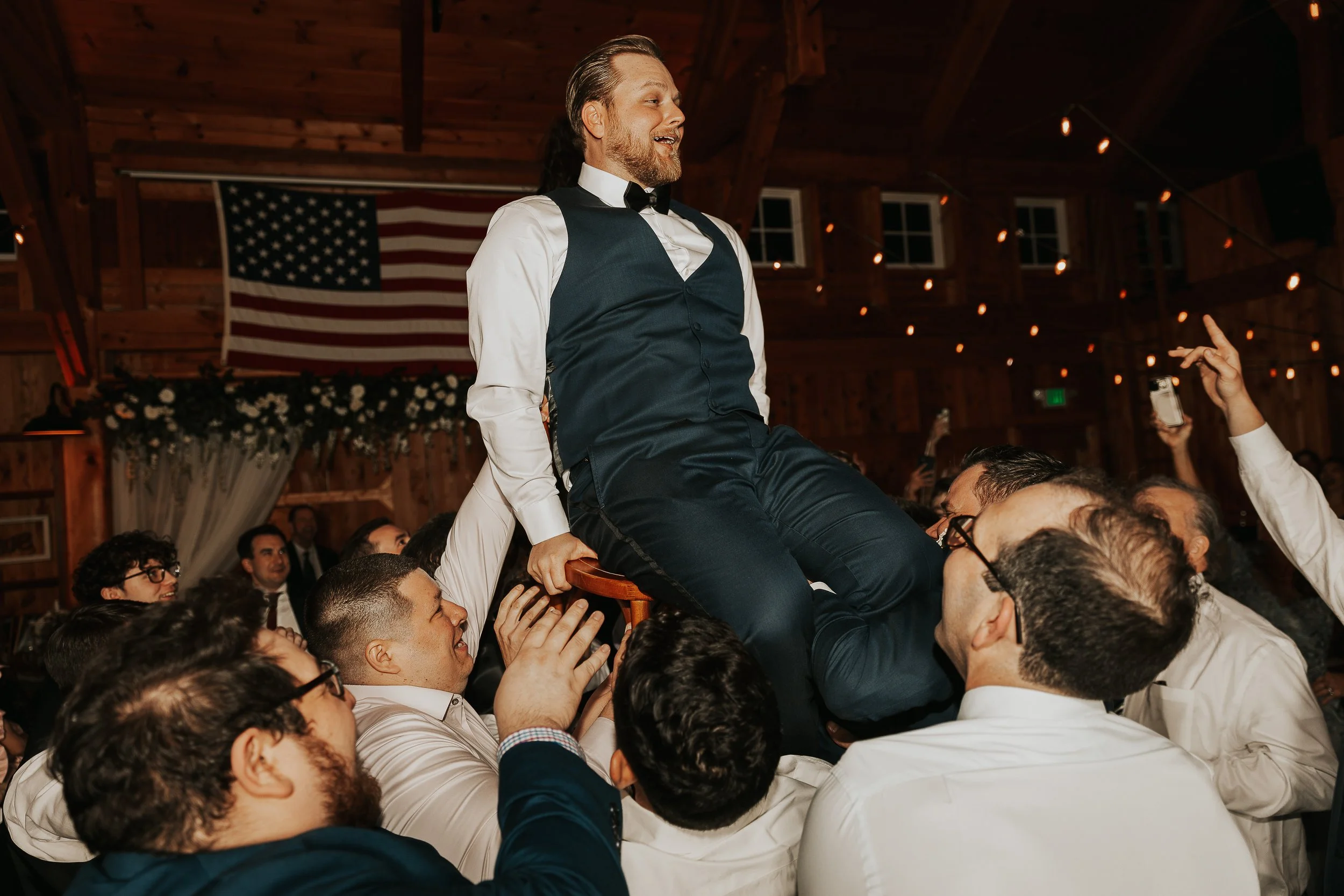A man with a beard wearing a suit and bow tie is sitting on a person's shoulders during a celebration or party. The indoor setting features wooden walls, string lights, and a large American flag in the background. Crowds of people are gathered around