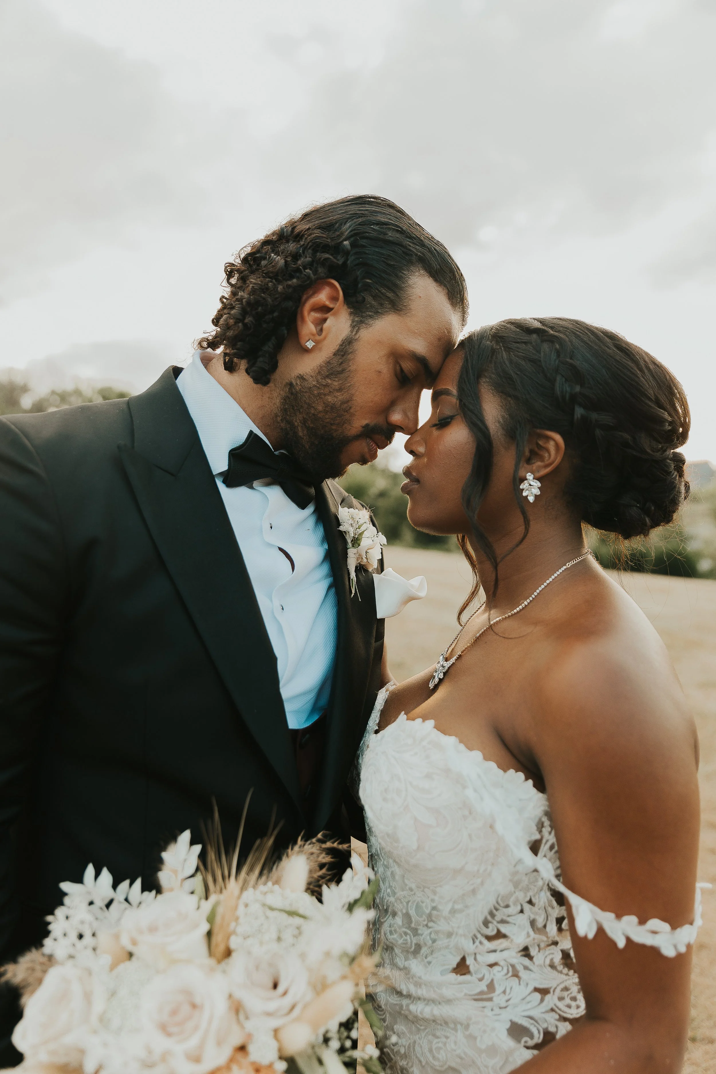 A bride and groom with foreheads touching, eyes closed, outdoors on a cloudy day, close-up shot of their faces and wedding attire, floral bouquet in foreground.