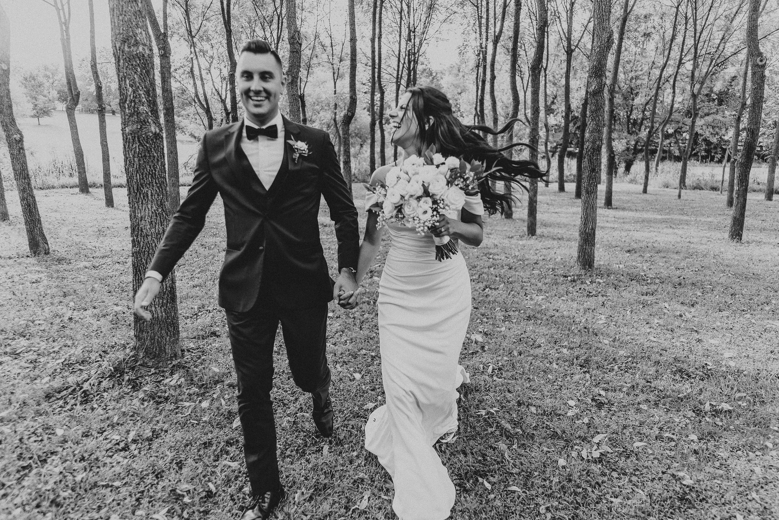 A black and white photo of a bride and groom walking hand in hand through a wooded area. The groom is wearing a tuxedo with a bow tie, and the bride is wearing a long wedding dress and holding a bouquet of flowers. Both are smiling and appear happy.