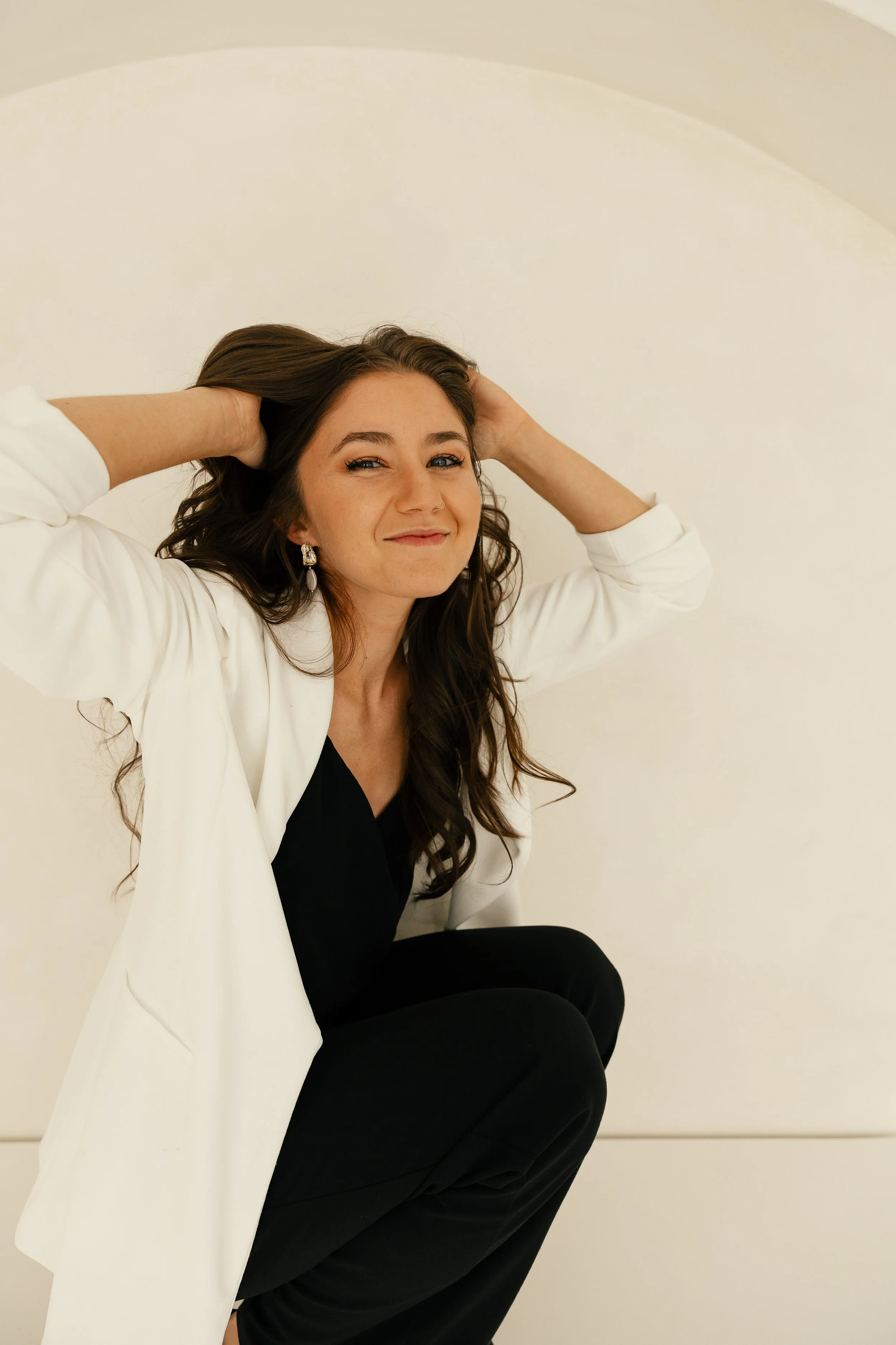 A photographer with long brown hair wearing a white blazer, black top, and earrings, sitting against a plain cream-colored wall, smiling slightly with her hands behind her head.