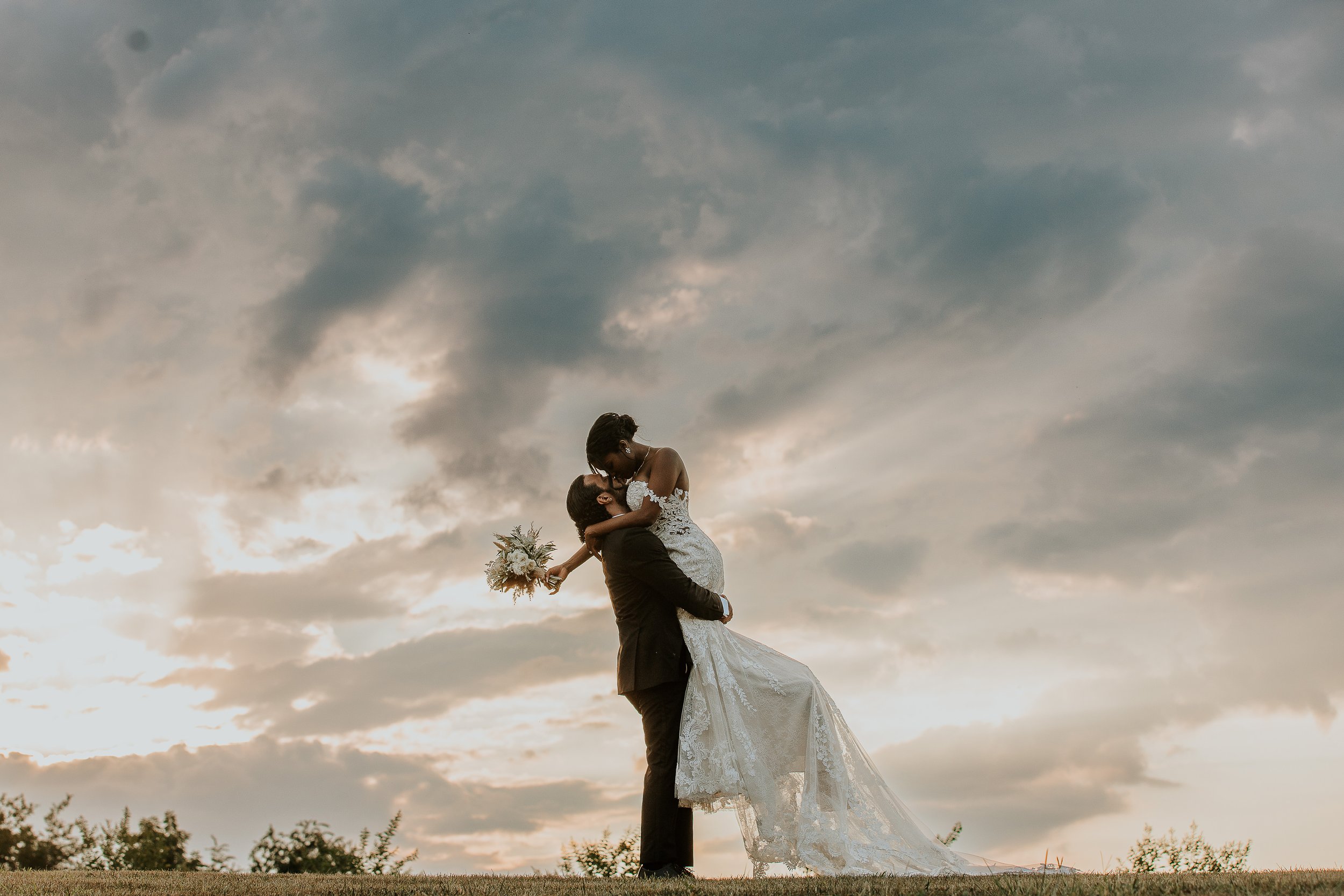 A couple in wedding attire, with the groom lifting the bride, against a cloudy sky at sunset.