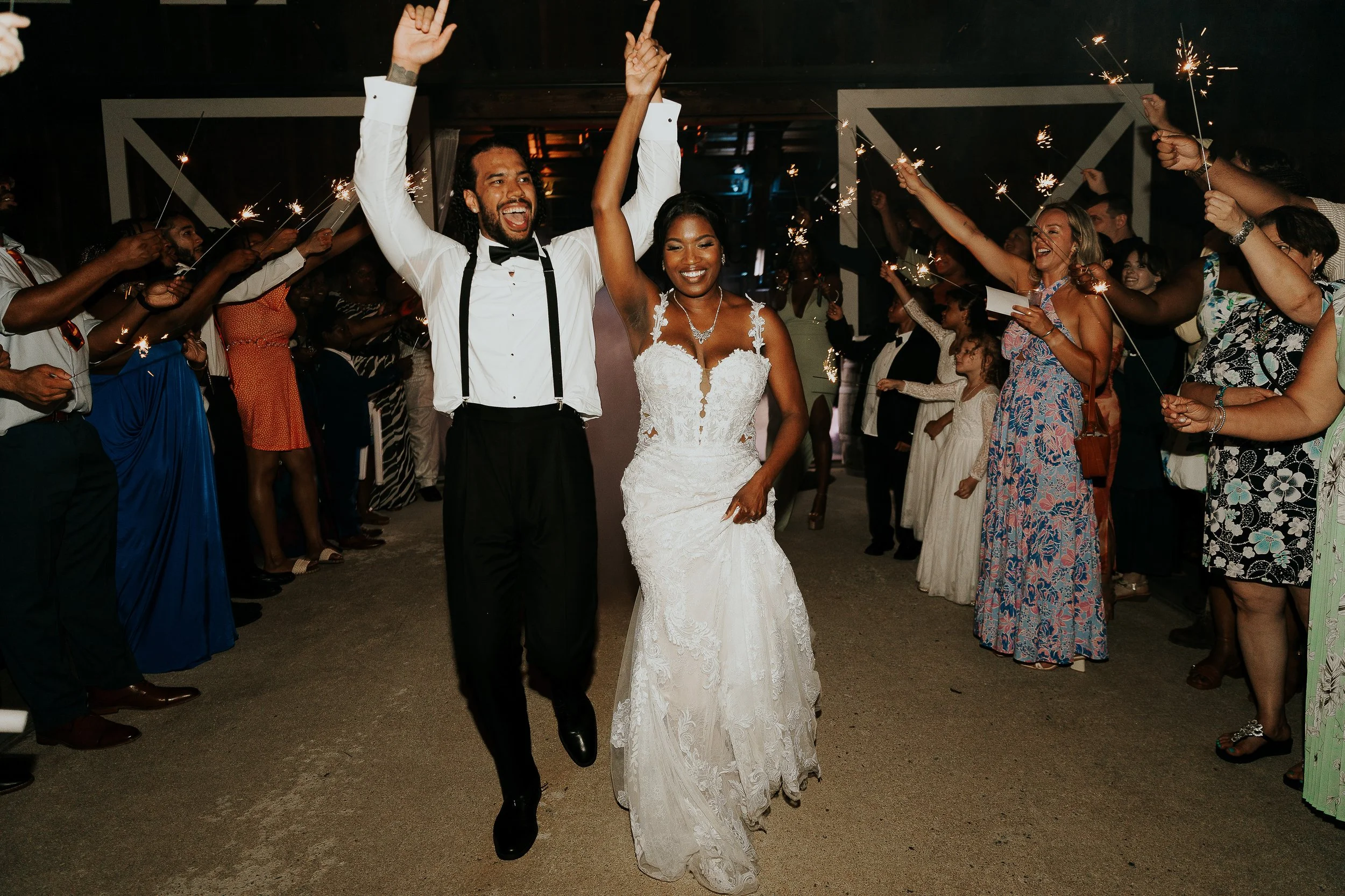 A newlywed couple, the groom wearing a tuxedo with suspenders and bow tie, and the bride in a lace wedding gown, celebrate with friends and family holding sparklers during a nighttime wedding reception.