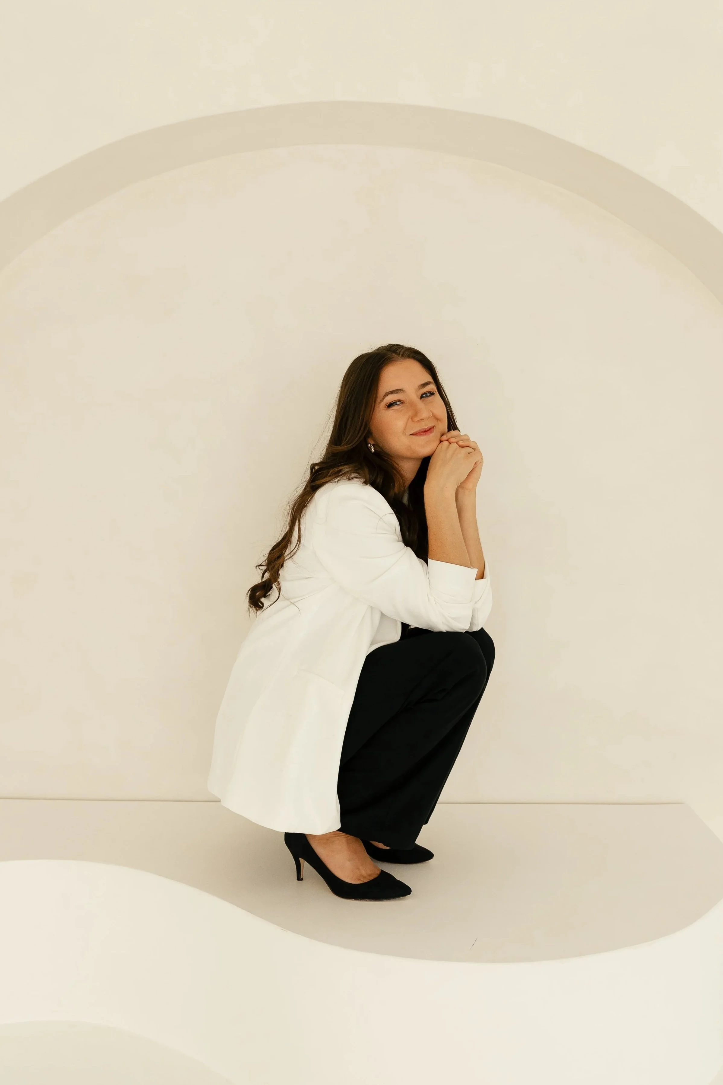 A photographer with long brown hair, wearing a white blazer, black pants, and black high heels, is squatting down with her hands clasped under her chin and smiling, against a plain off-white background with a faint arch shape.
