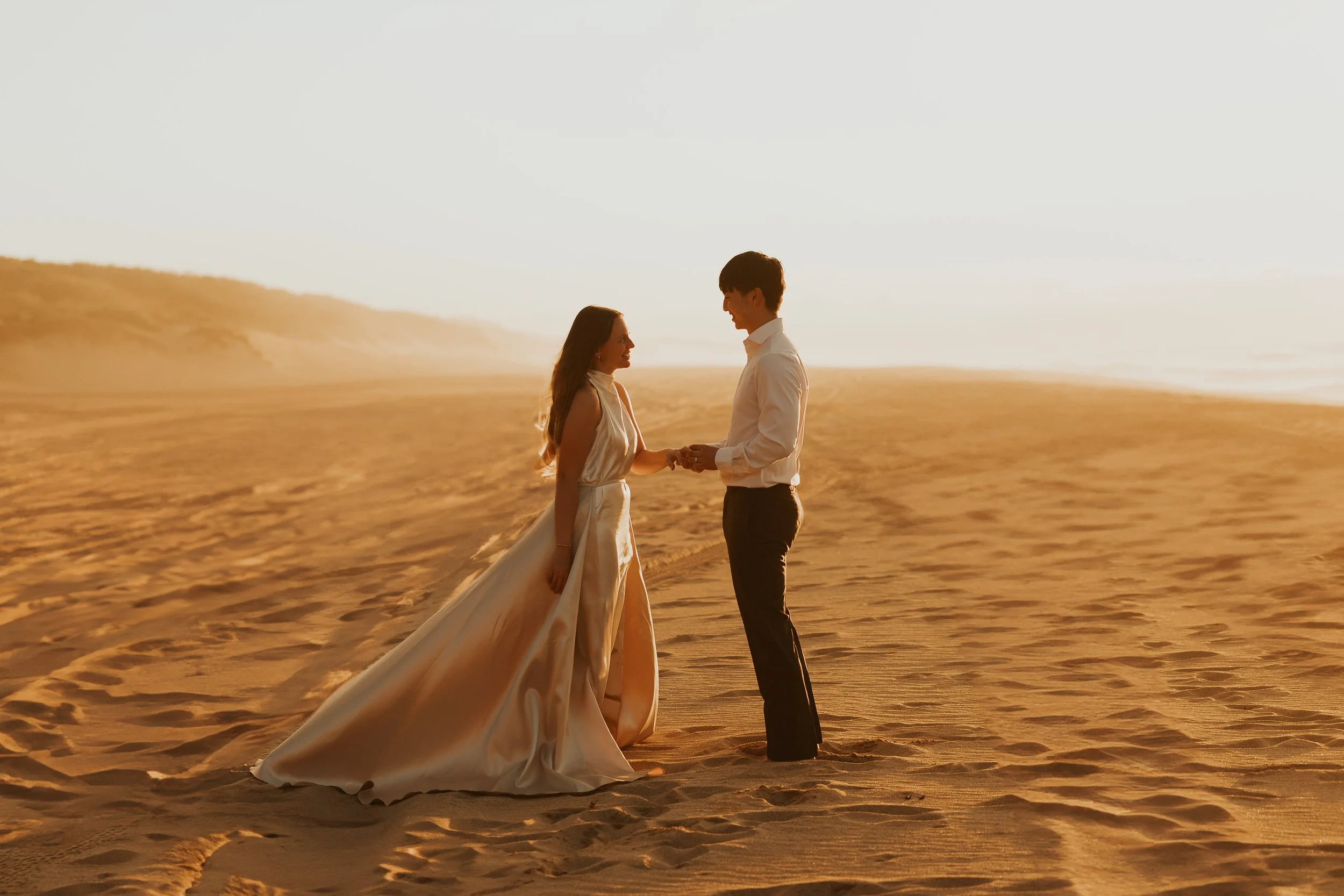 A destination wedding couple holding hands on a sandy beach during sunset, dressed in formal attire, with the woman wearing a white gown and the man in black pants and a white shirt.
