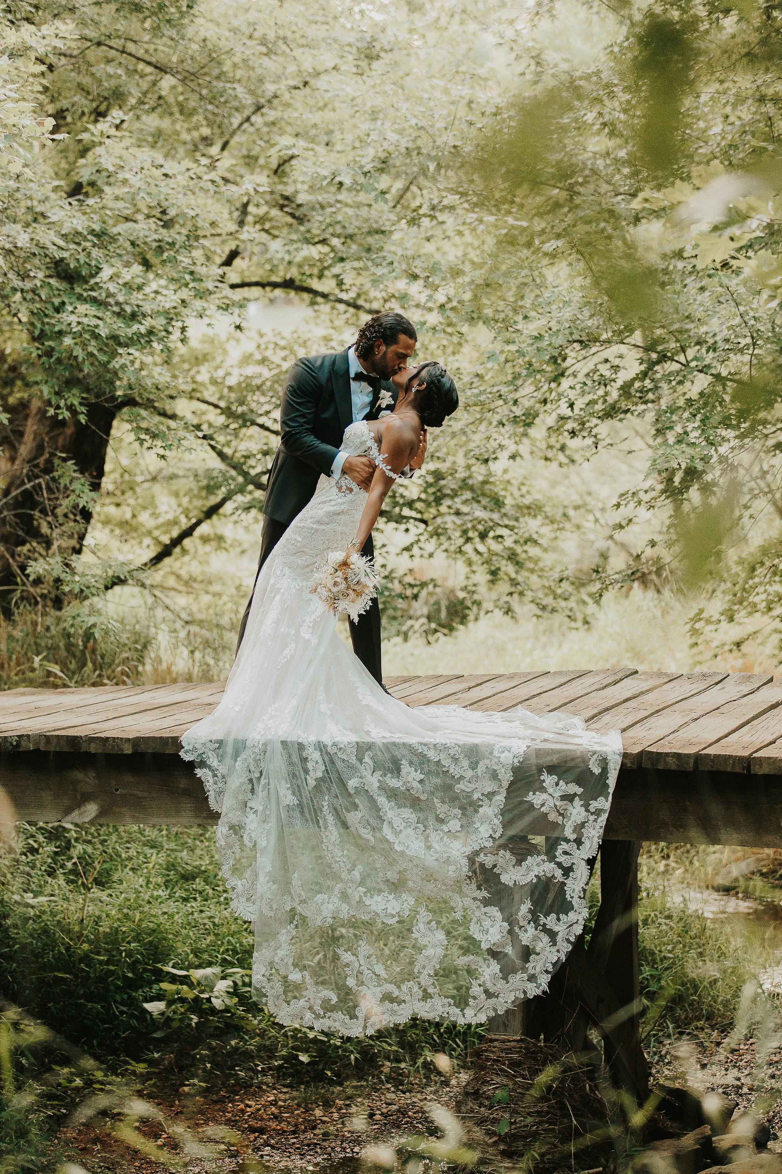 A wedding couple kissing on a wooden bridge surrounded by lush green trees.