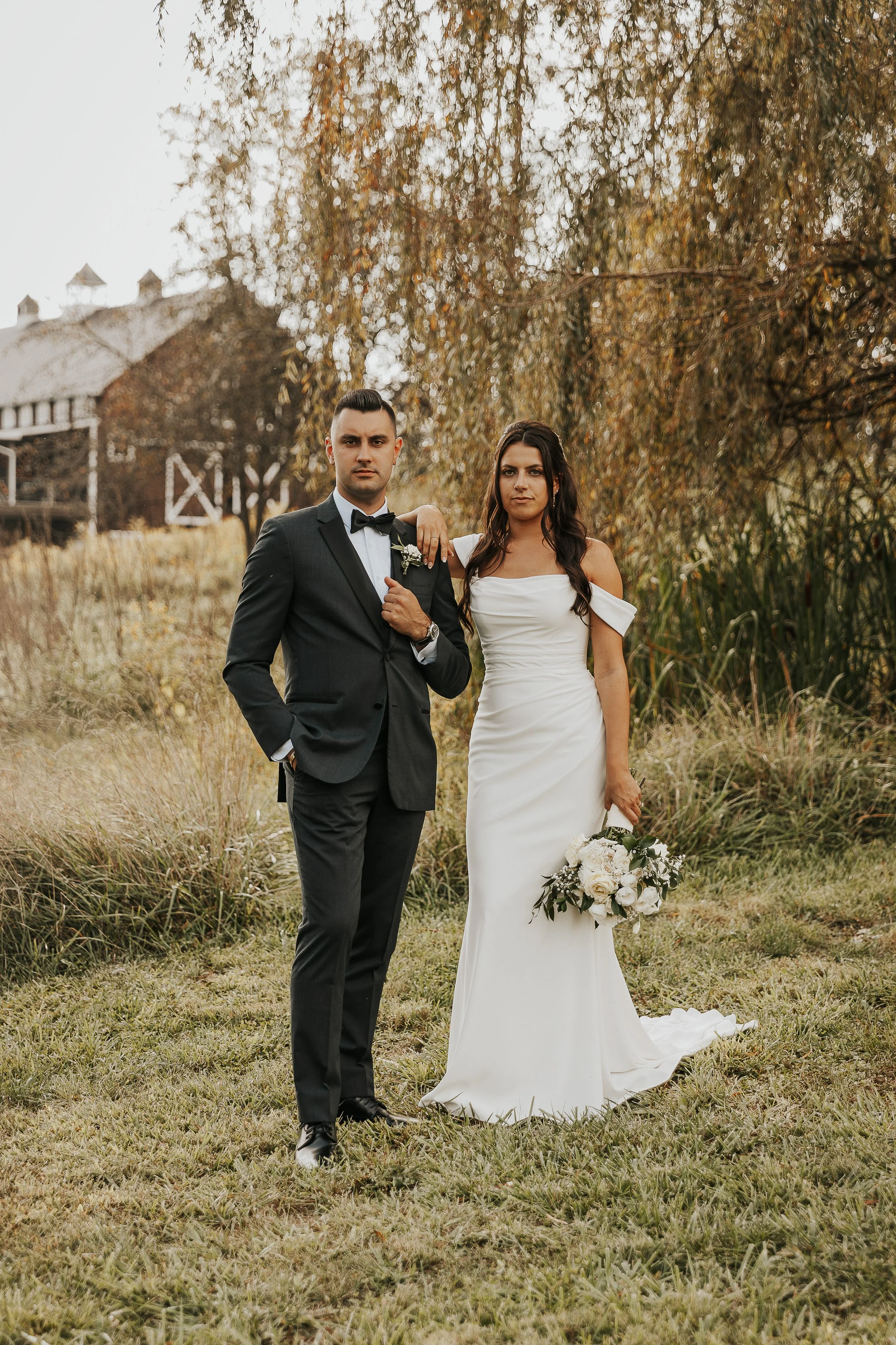 A bride and groom in formal wedding attire standing outdoors in a grassy area with autumn trees and a barn in the background.