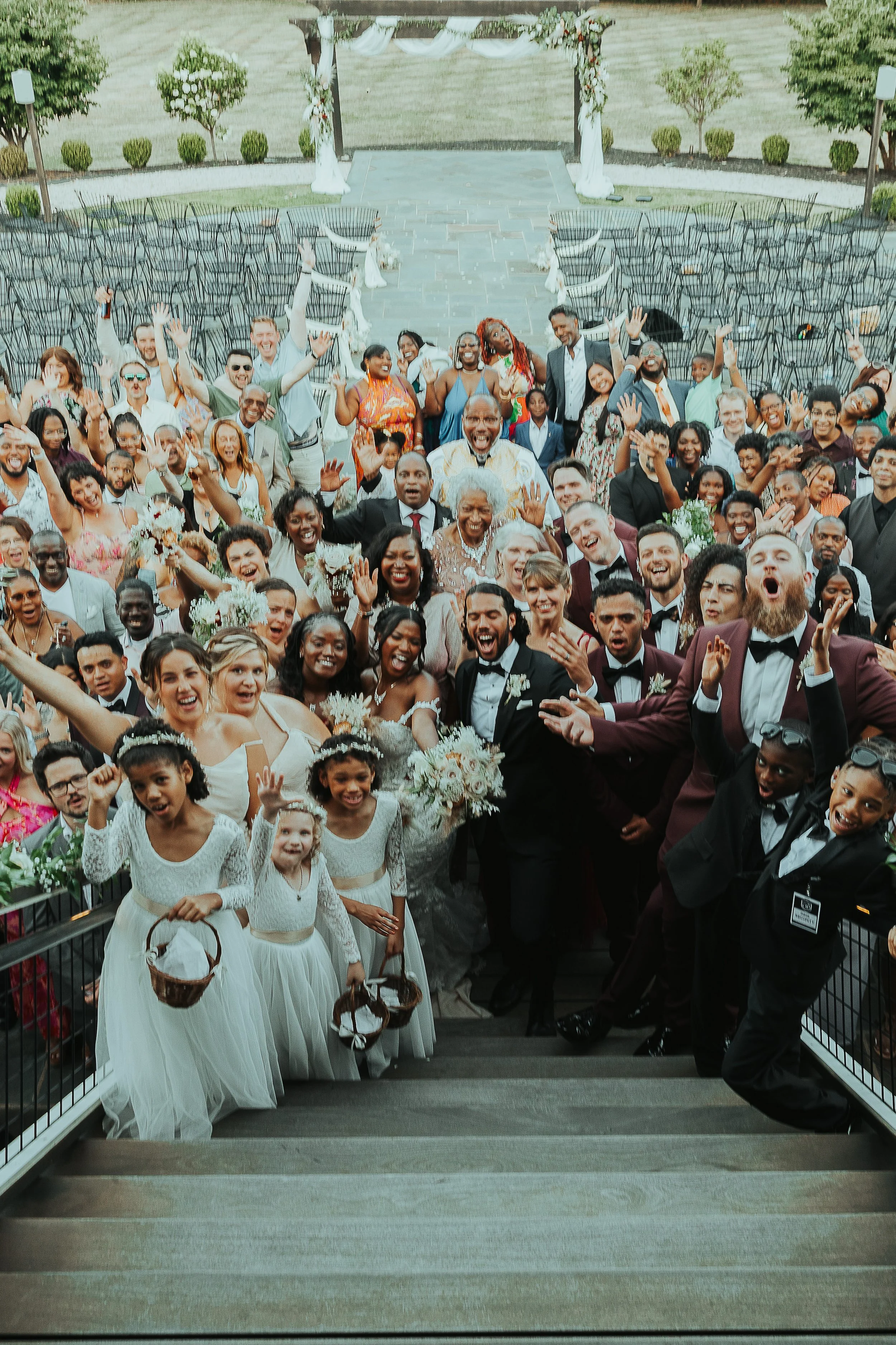 A large group of wedding guests gathered at the bottom of a staircase, smiling and waving, with a wedding ceremony area set up in the background decorated with flowers and white drapes.