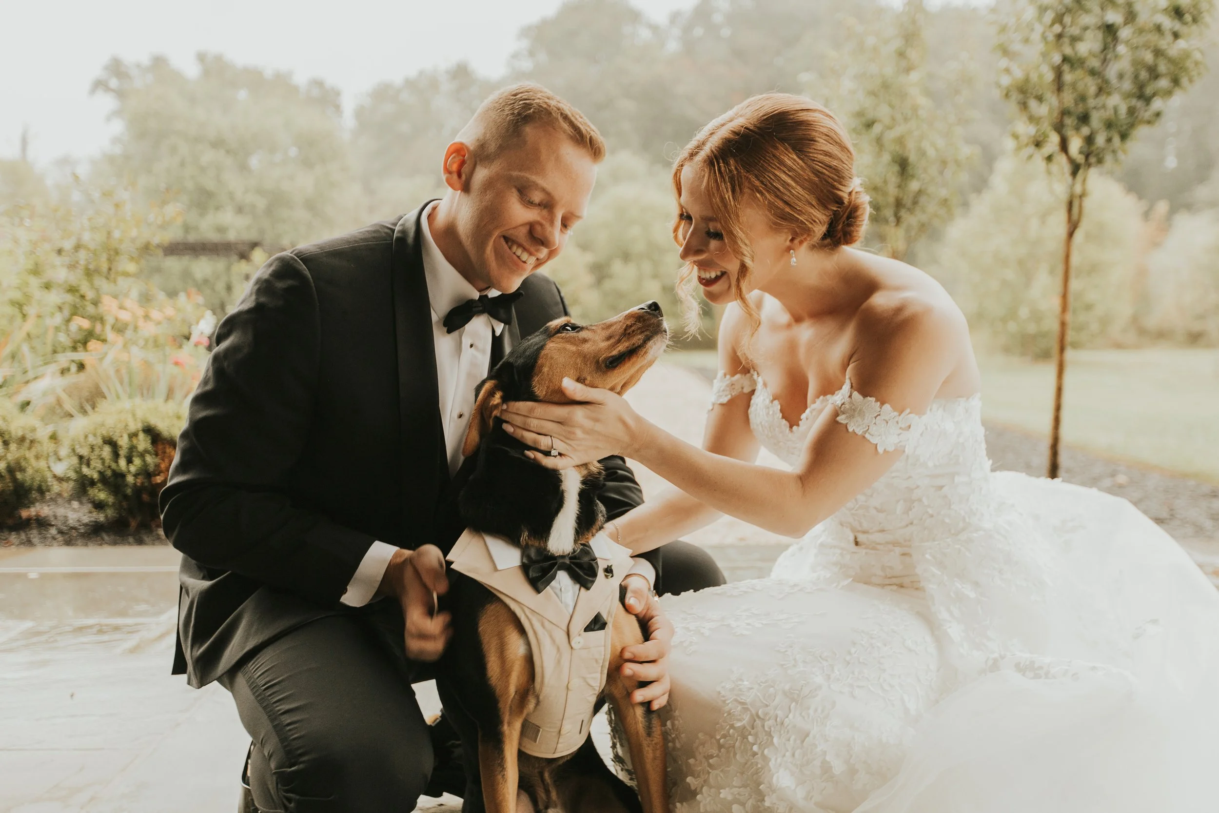 A bride and groom in wedding attire smiling and interacting with a large dog, who is dressed in a tuxedo and sitting between them outdoors near a body of water, with trees and greenery in the background.