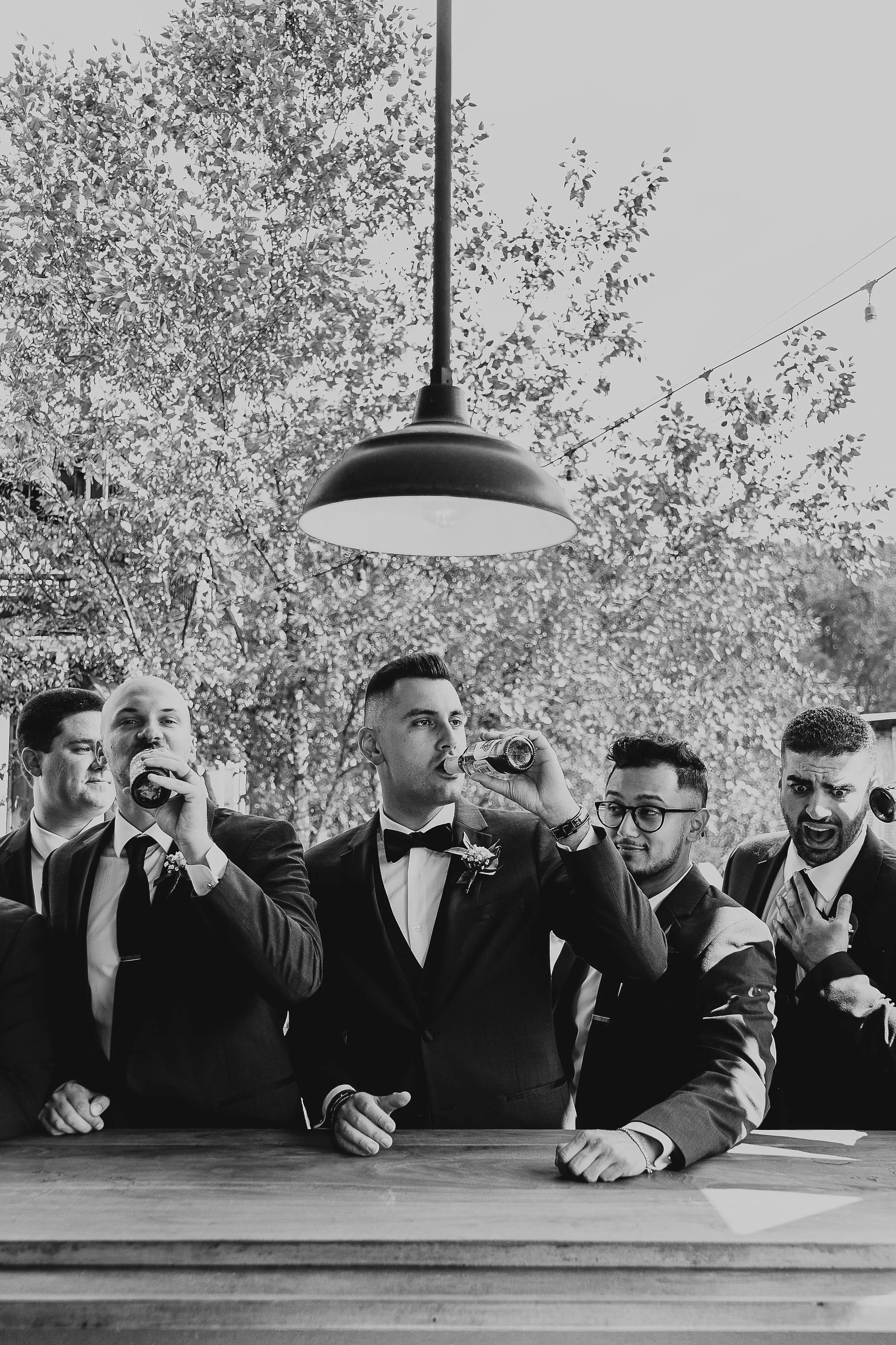 A group of men dressed in suits and ties, some holding drinks, gathered around a wooden table outdoors with trees and string lights overhead.