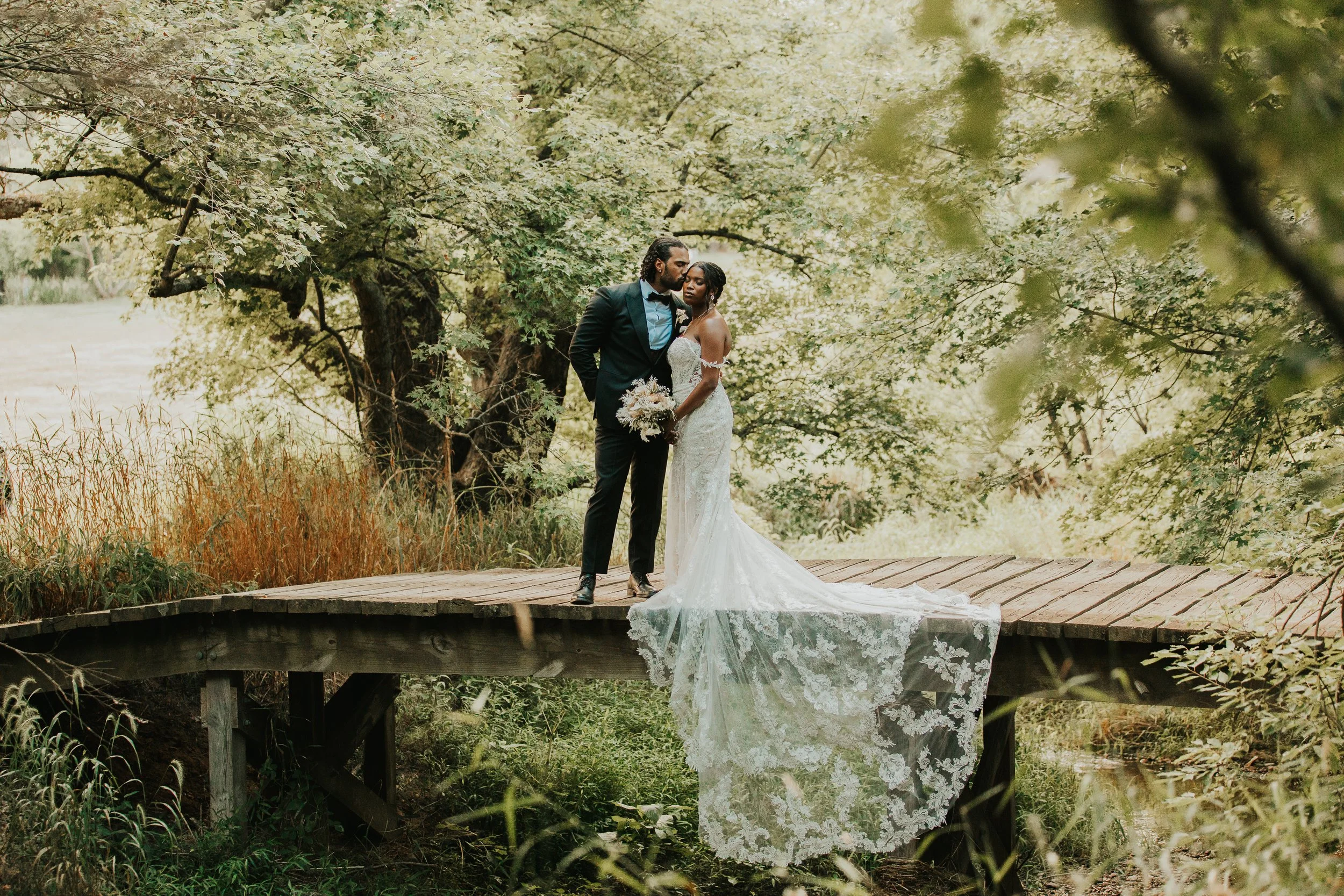A bride and groom standing on a wooden bridge surrounded by lush green trees, with the bride holding a bouquet and wearing a white lace wedding dress.