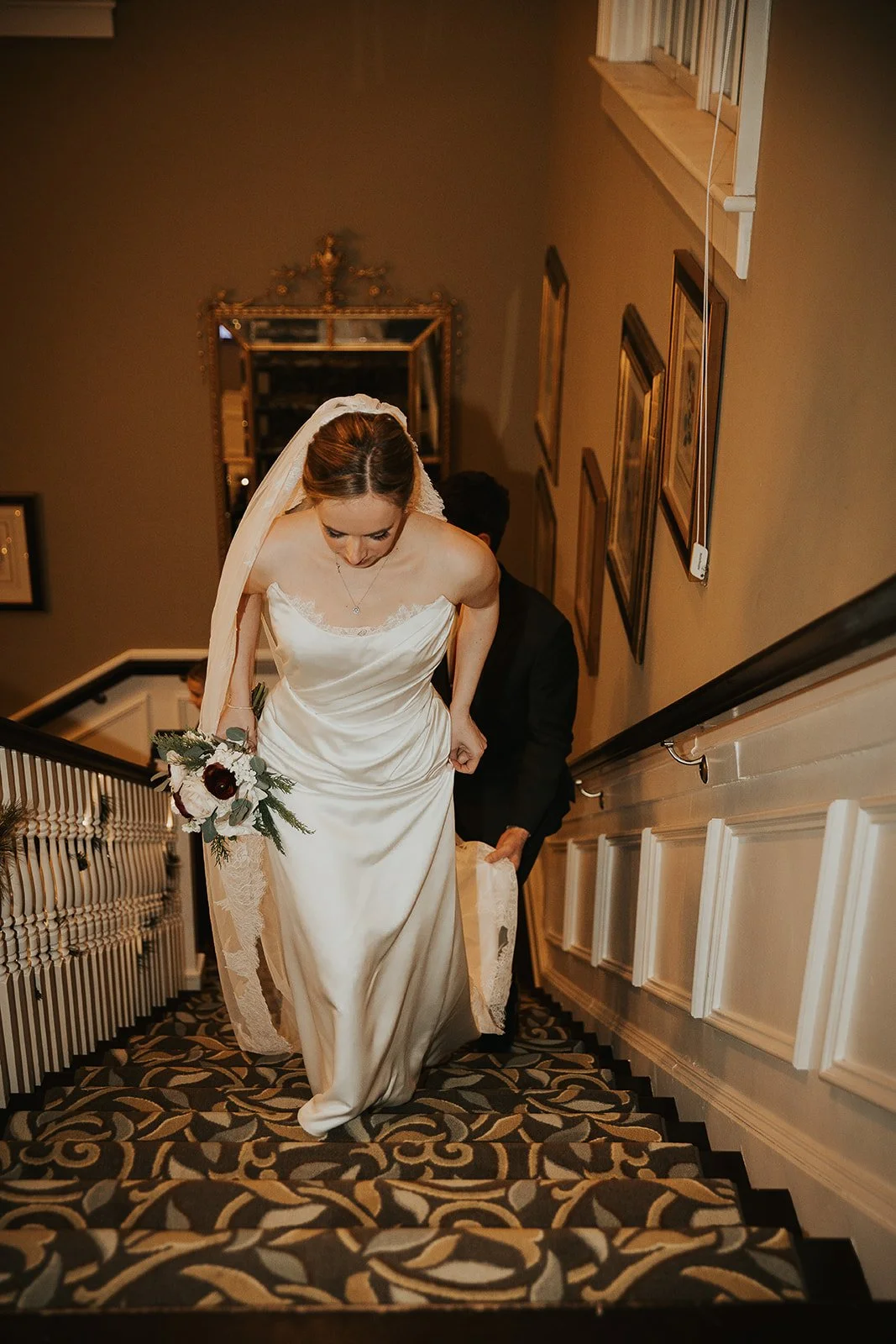 A bride in a white wedding dress holding a bouquet as she ascends a staircase, with a man behind her helping lift her gown.