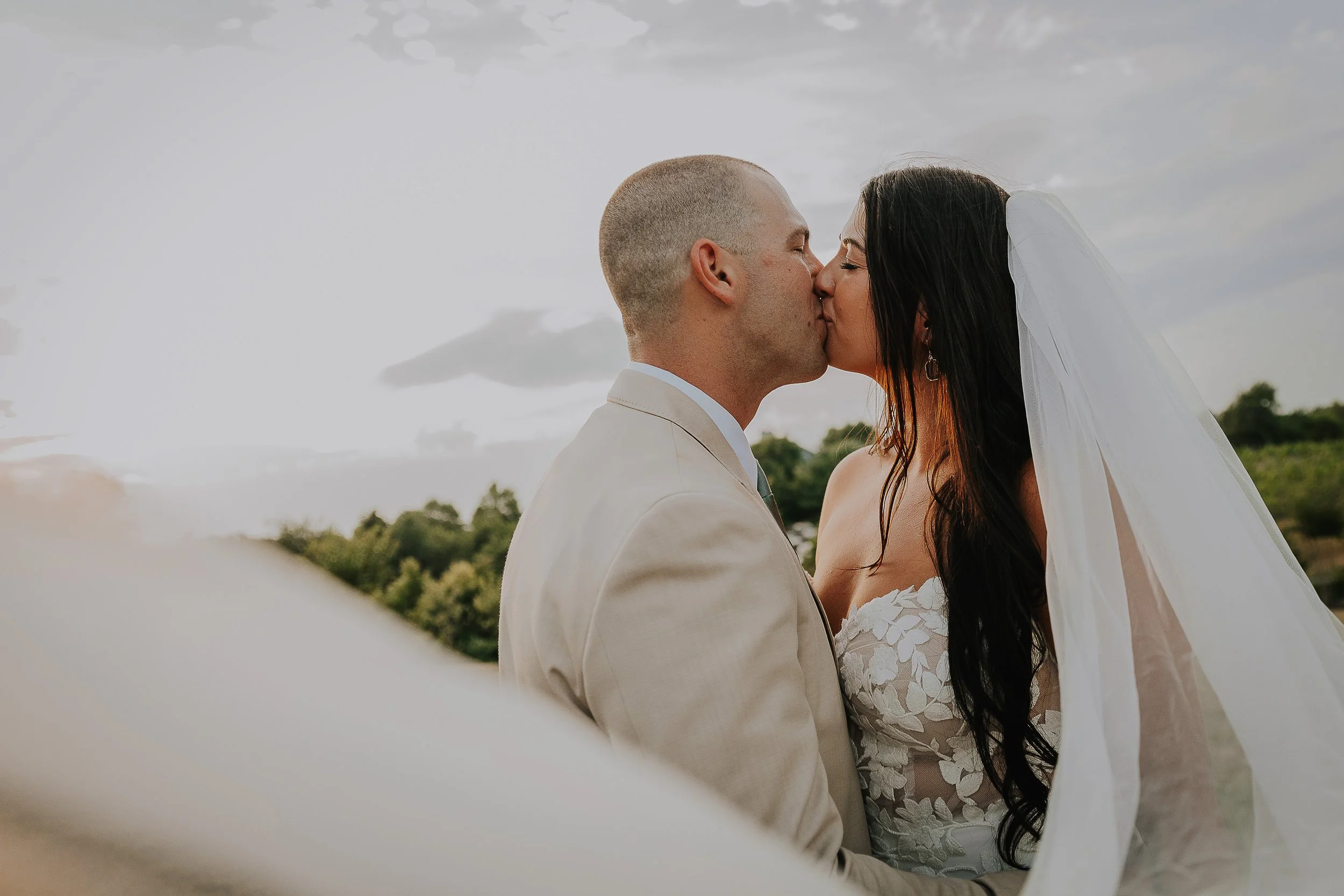 A bride and groom kissing outdoors during their wedding, with a cloudy sky and greenery in the background.