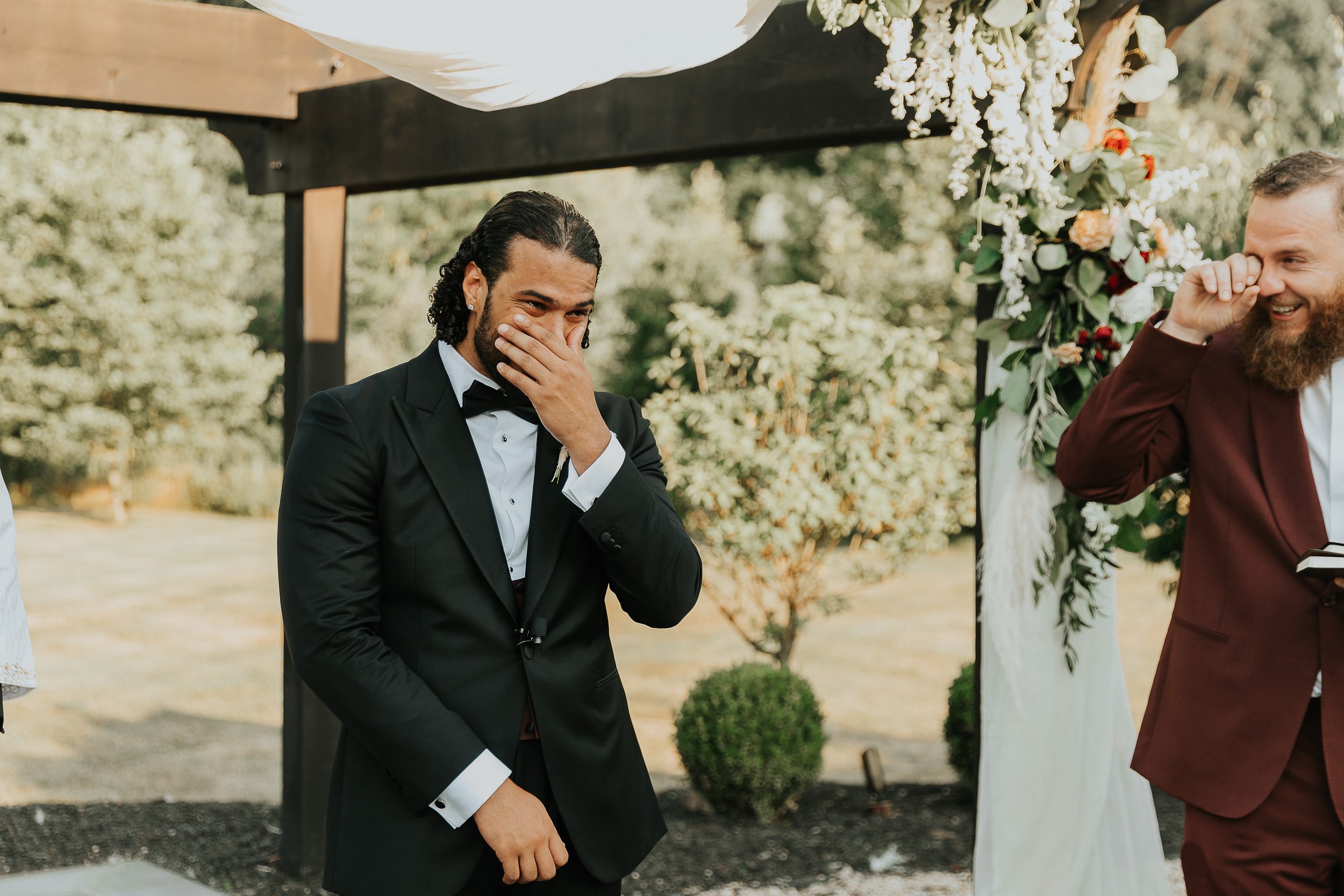A groom in a black tuxedo with a bow tie is smiling and covering his mouth, standing outdoors during a wedding ceremony. another man in a maroon suit is smiling and wiping his eye, standing beside a decorated wedding arch with flowers, in a natural o