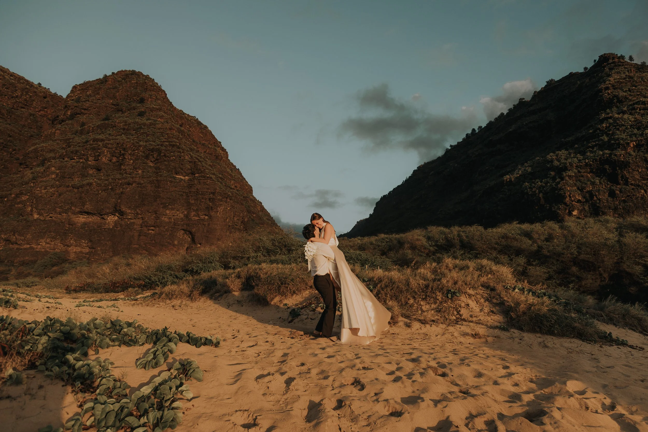 Destination wedding couple dressed in wedding attire sharing a kiss in a desert landscape with large mountains in the background during sunset.