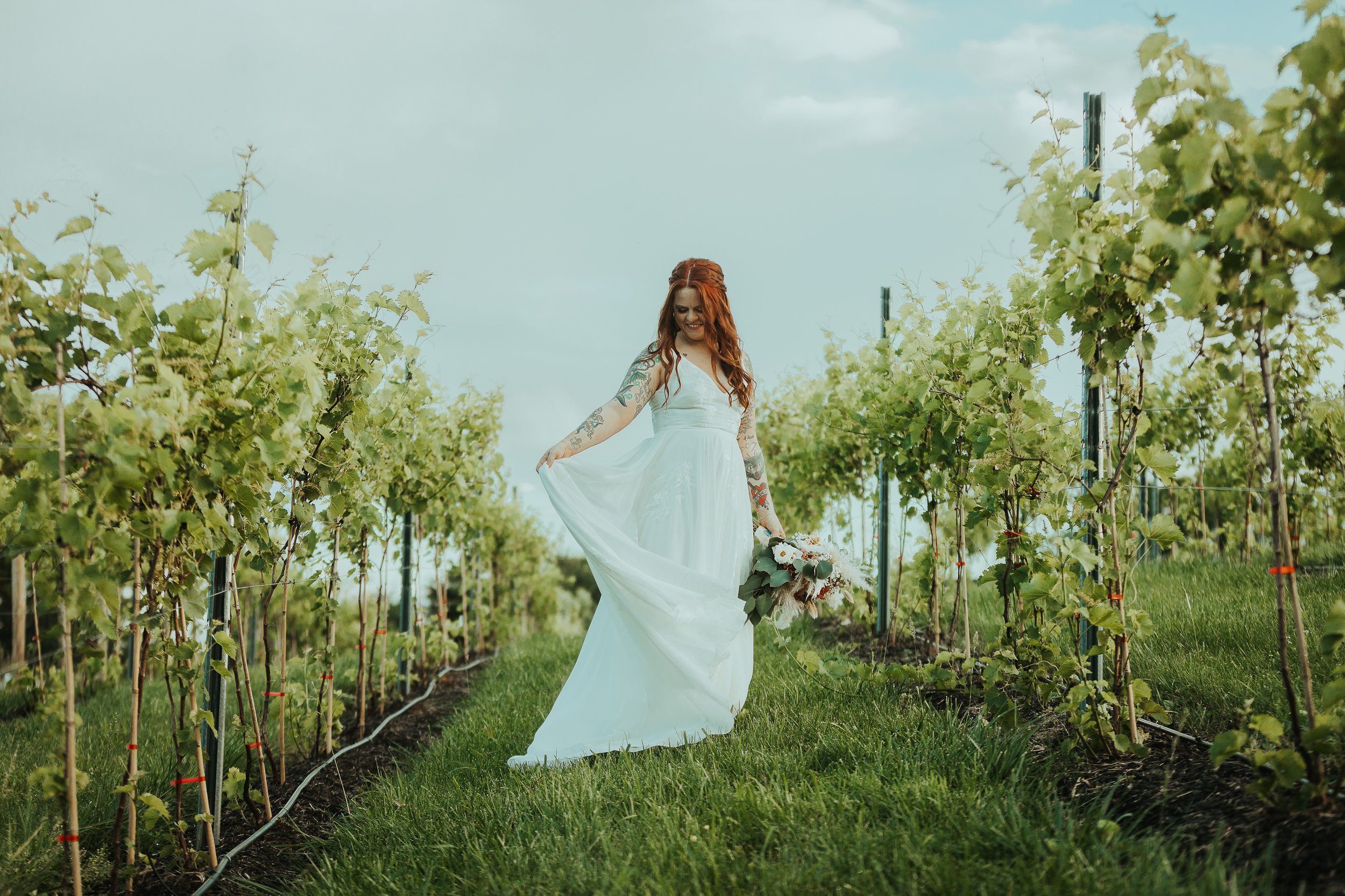 A woman with red hair wearing a white wedding dress walking through a vineyard holding a bouquet of flowers.