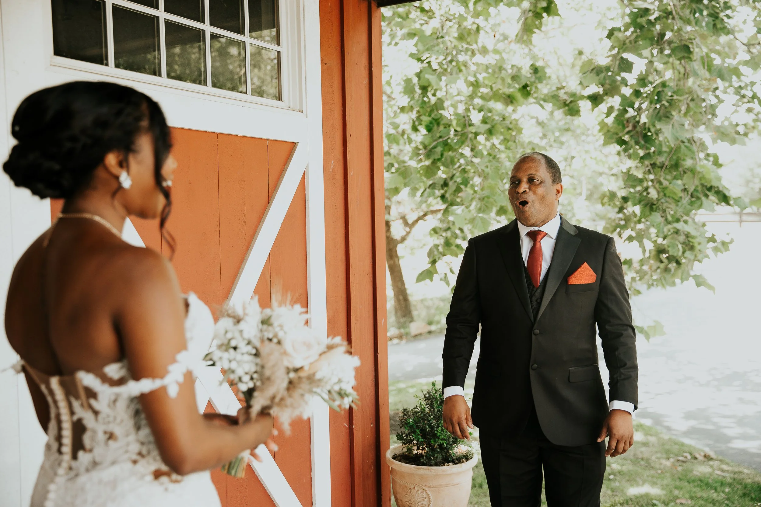 A man dressed in a black suit with a red tie and pocket square is standing outside, looking surprised or excited, in front of a woman holding a bouquet of white flowers, with a red building and green trees in the background.