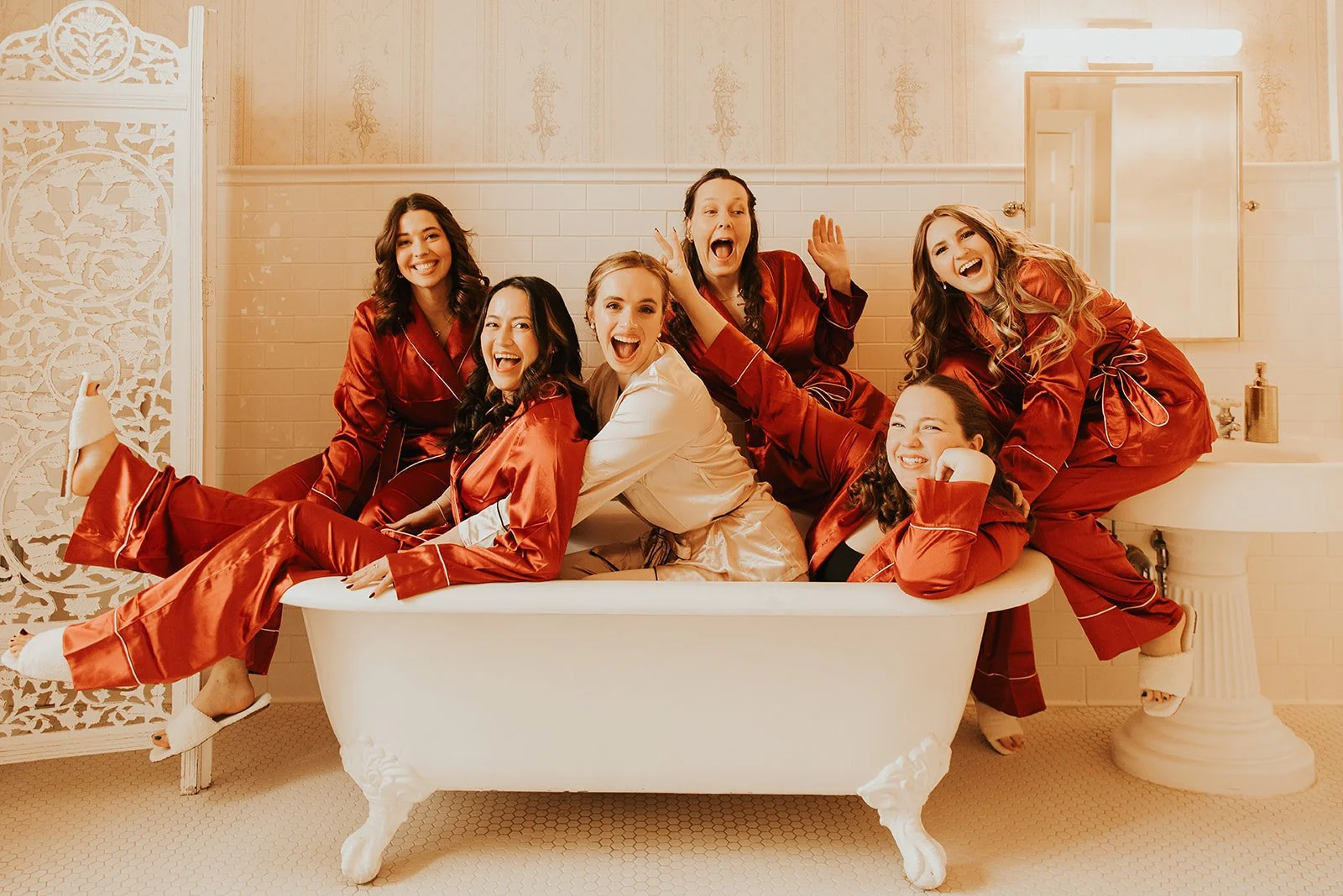 Group of seven women in matching satin pajamas sitting and laying in an old-fashioned bathtub, smiling and laughing in a warmly lit bathroom.