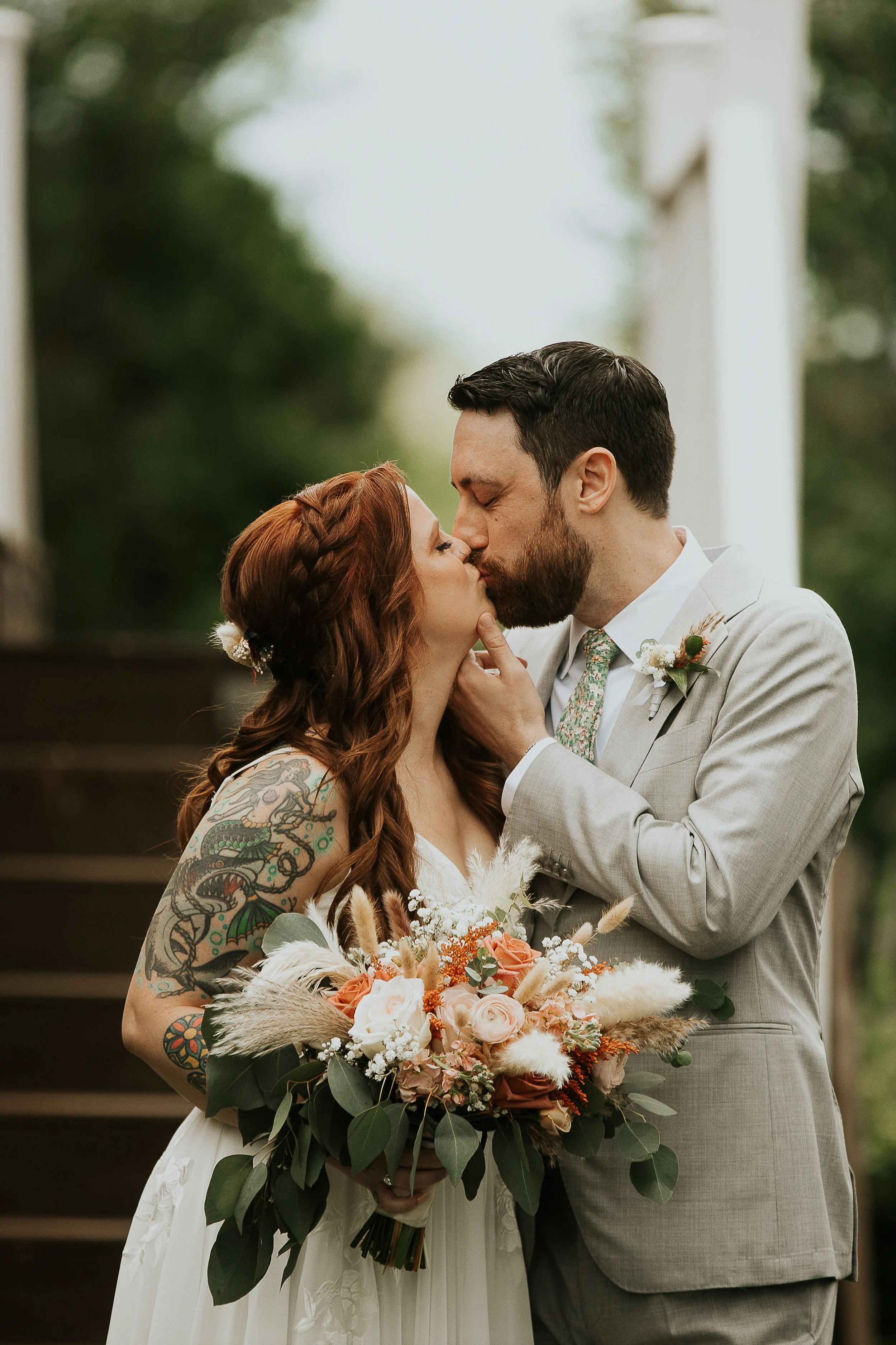 An elopement couple getting married, sharing a kiss during the ceremony. The bride has tattoos on her arm and is holding a bouquet of flowers, while the groom is in a light gray suit and boutonniere. The background is blurred greenery.