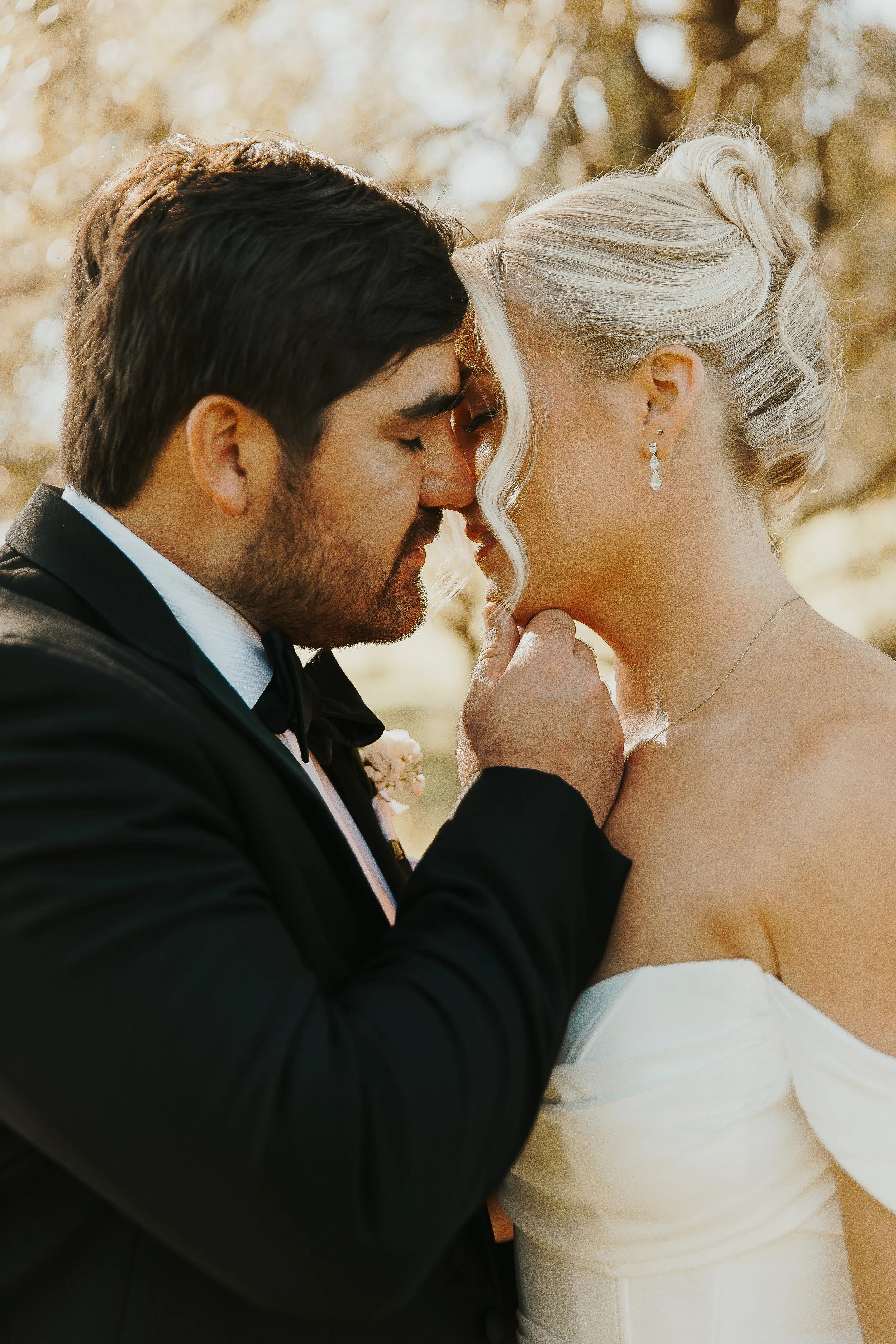 A man and woman, dressed in wedding attire, are sharing an intimate moment with foreheads touching and eyes closed, outdoors with a blurred background of sunlight and autumn trees.