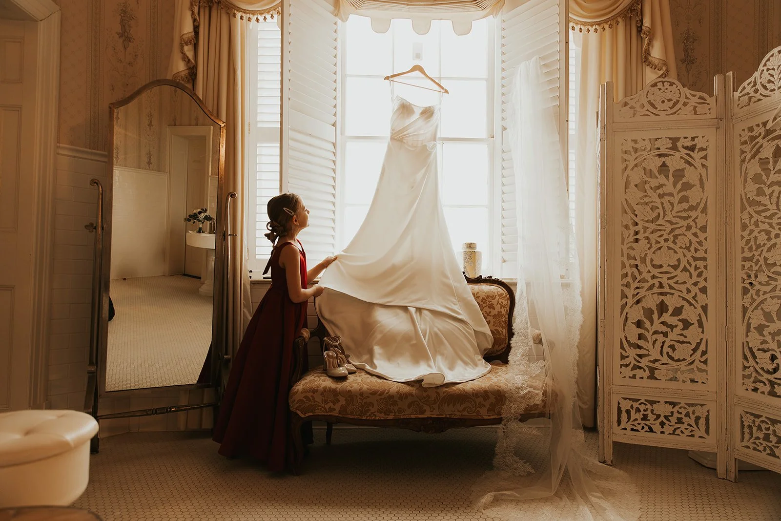 A young girl in a red dress looking at a wedding dress hanging in front of a window in a vintage, well-lit room with ornate curtains and wallpaper.
