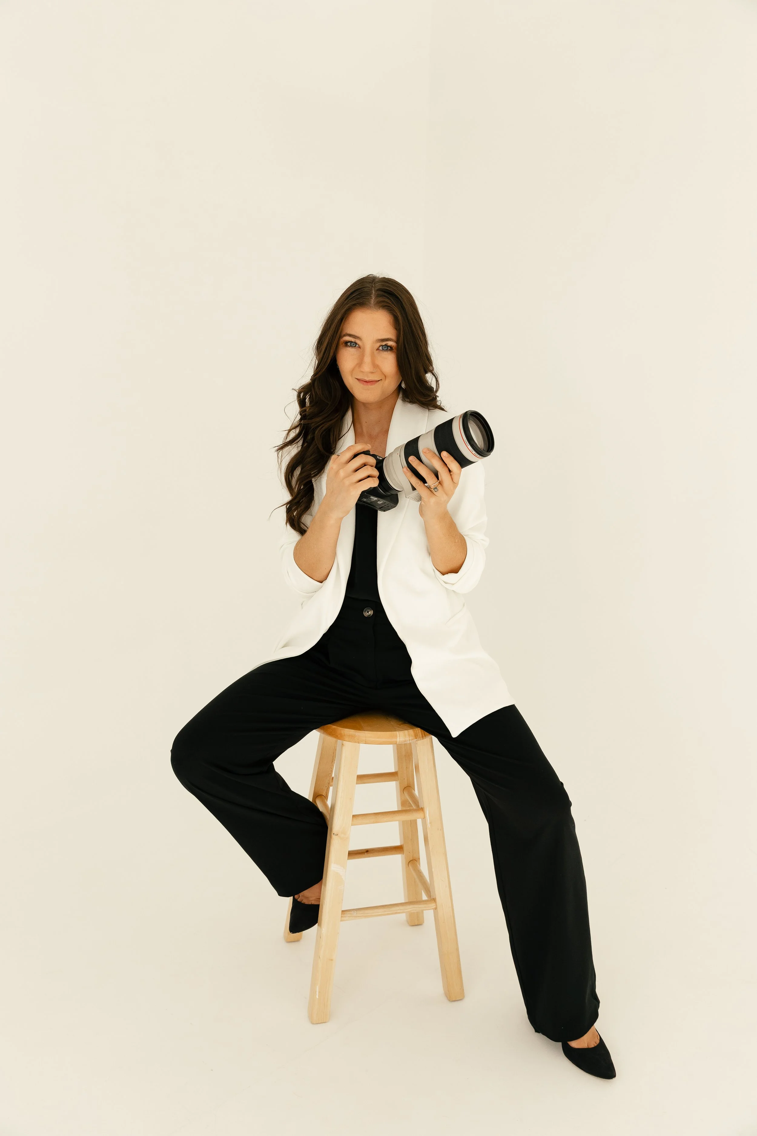 A photographer with long brown hair wearing a white blazer, black pants, and black shoes, sitting on a wooden stool with a landscape camera in her hands against a plain light background.