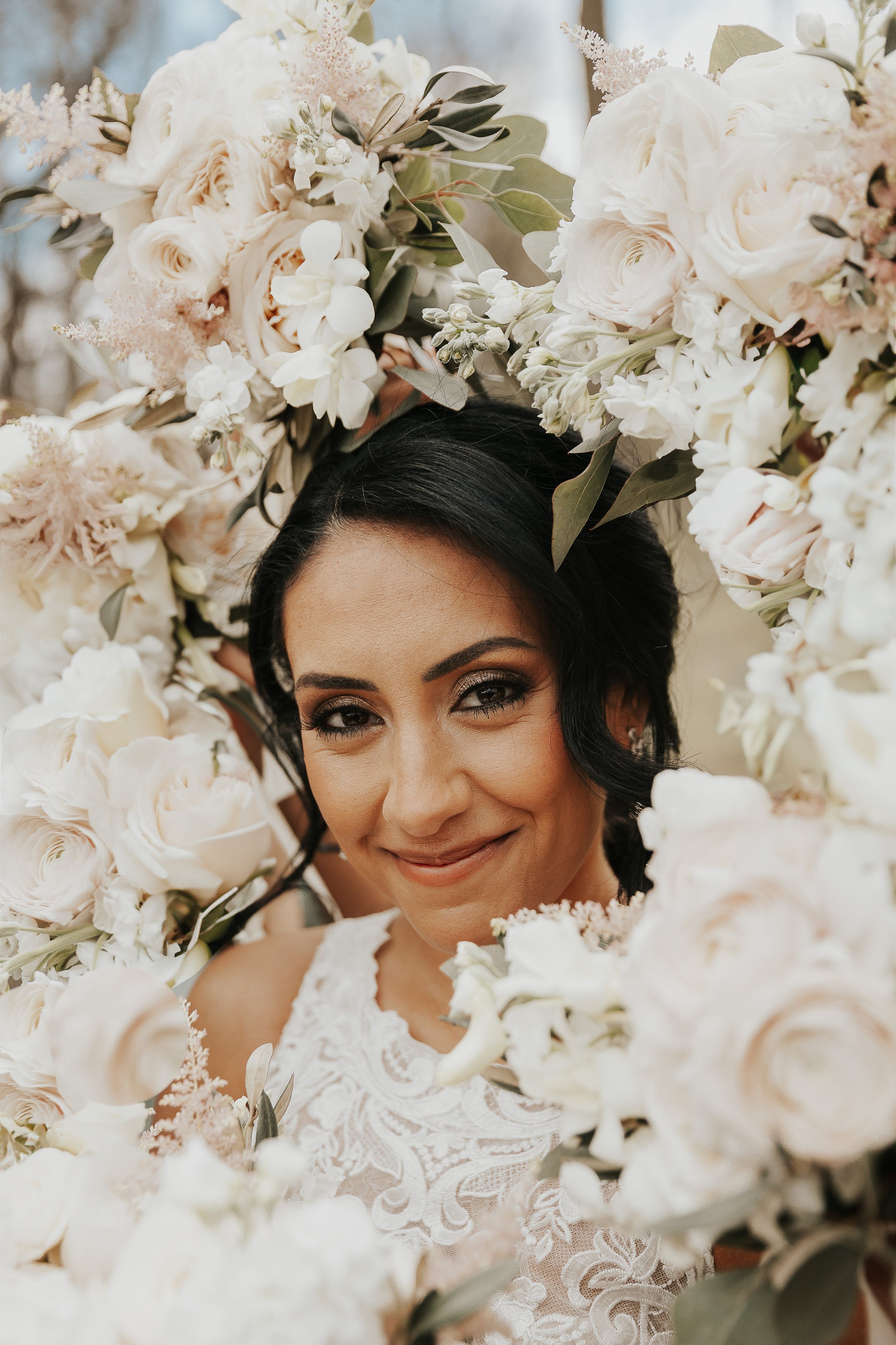 A woman with dark hair and makeup smiling, surrounded by white roses and other flowers, with part of her face and upper body visible.