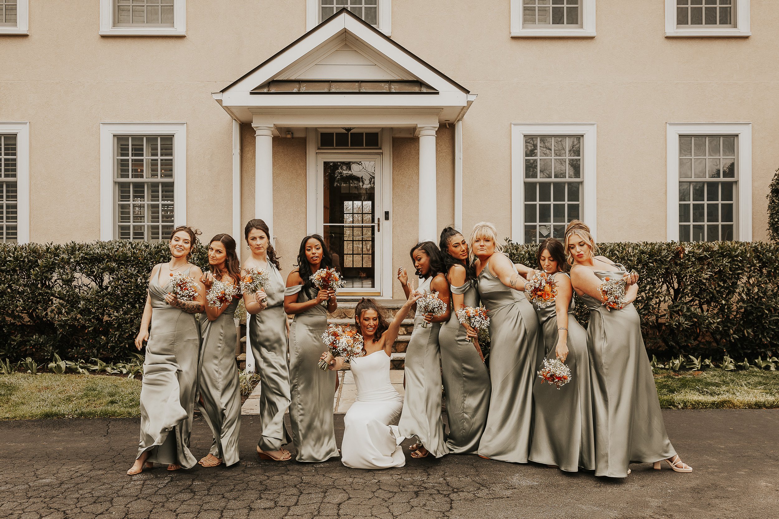 Group of nine women in gray satin dresses standing on steps in front of a cream-colored house with white trim, holding bouquets of flowers, celebrating at a wedding.