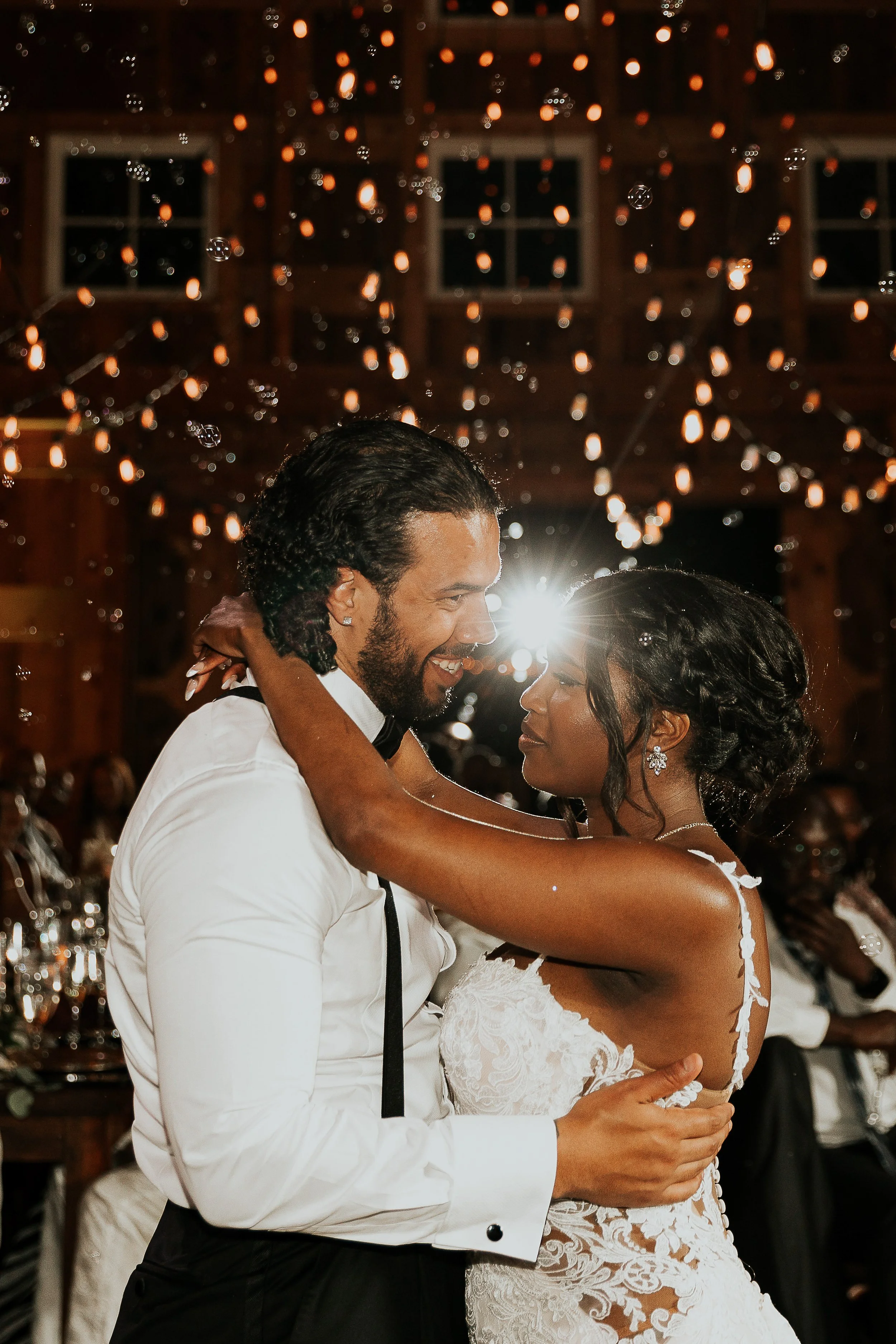 A couple dancing at a wedding reception with warm lighting and wooden ceiling, the groom in a tuxedo and the bride in a lace wedding dress.