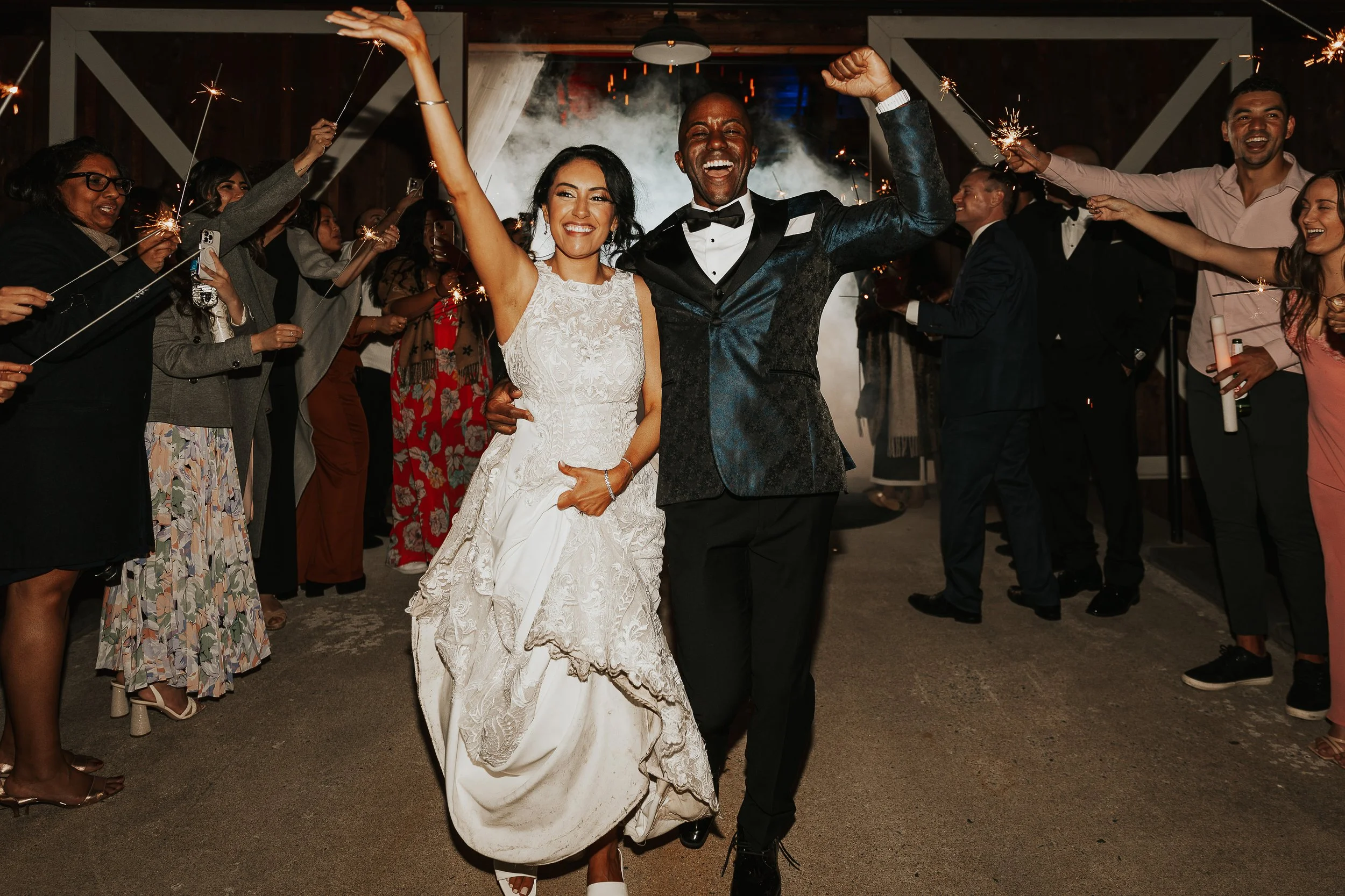 Couple celebrating at their wedding reception with sparklers, surrounded by friends, in an indoor venue.