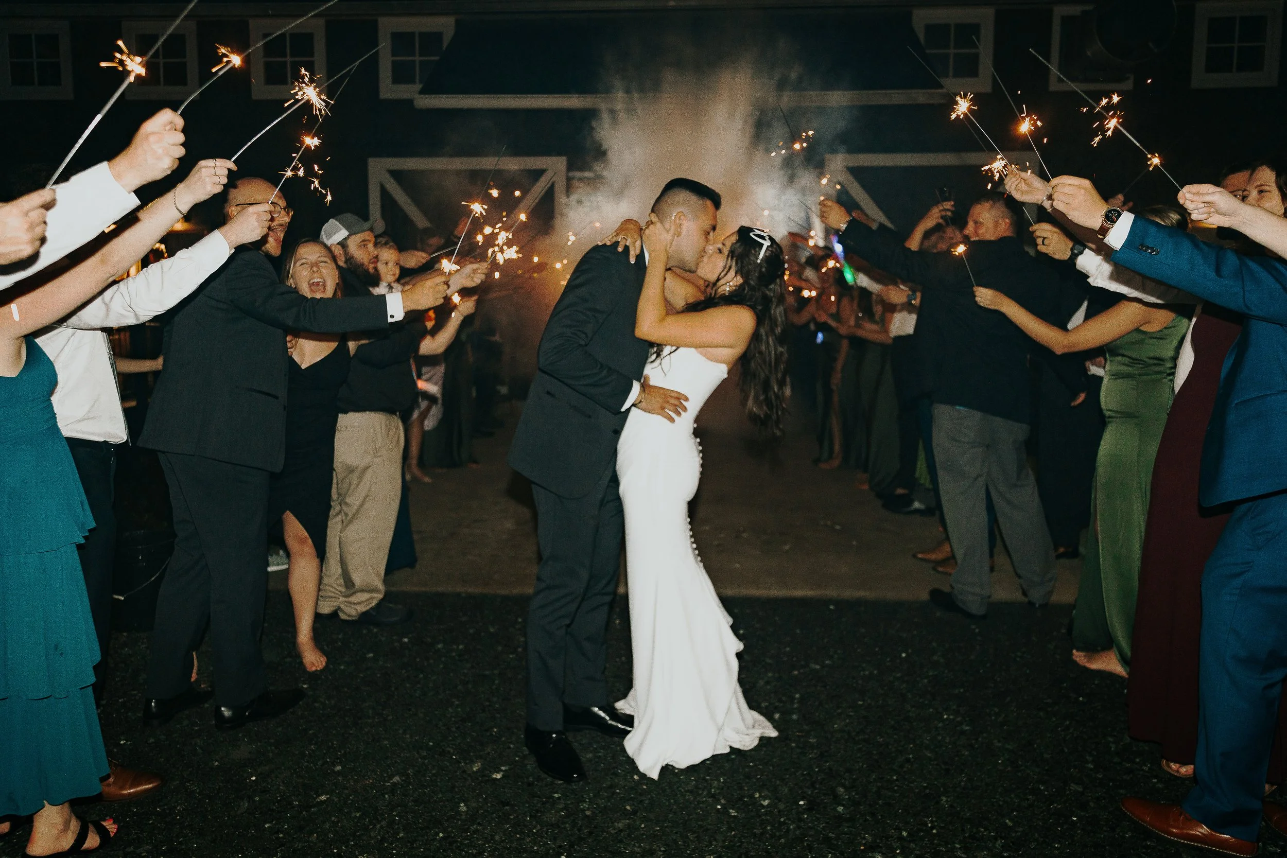A newlywed couple sharing a kiss at night surrounded by guests holding sparklers.