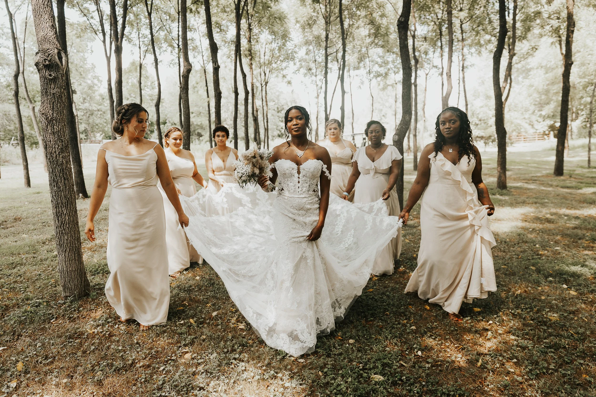 A bride in a white lace wedding dress holding a bouquet, walking through a wooded area with five bridesmaids in cream dresses.