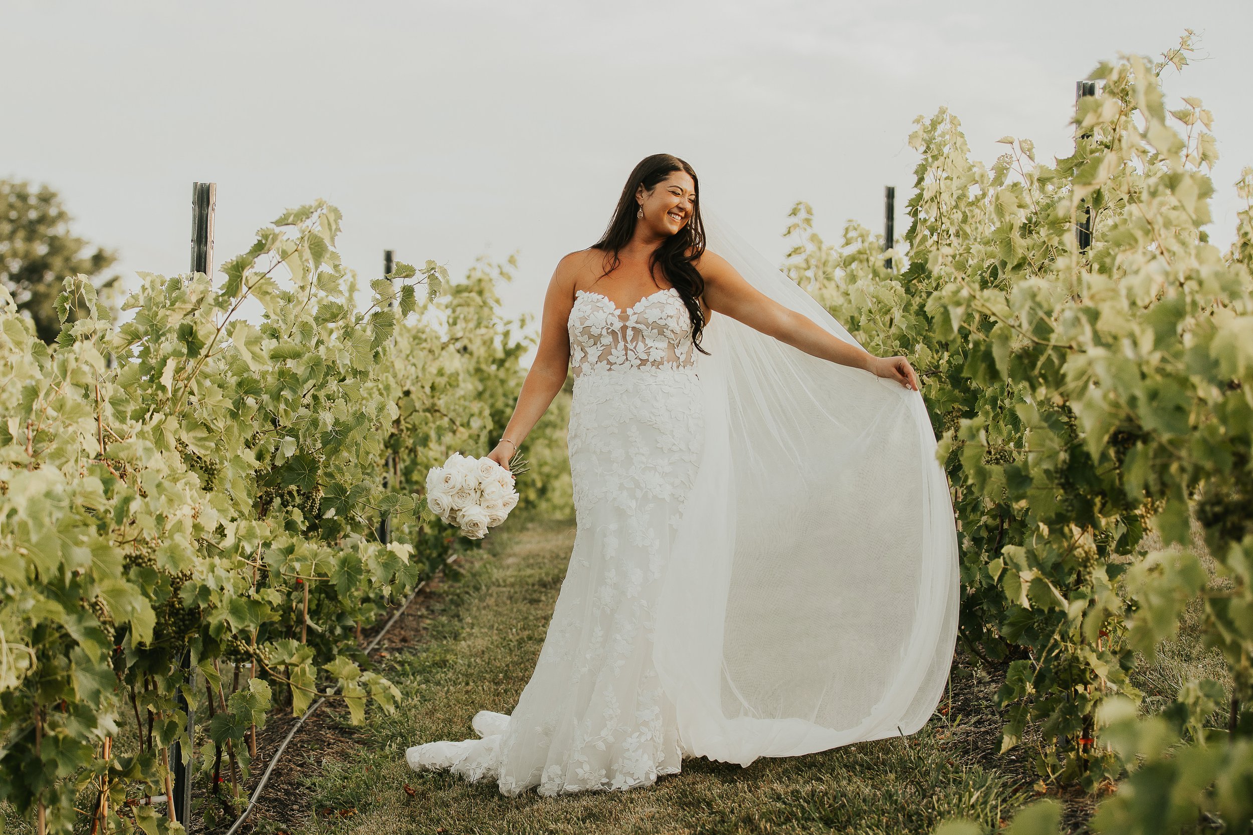 A woman in a wedding dress smiling in a vineyard holding a bouquet of white roses.