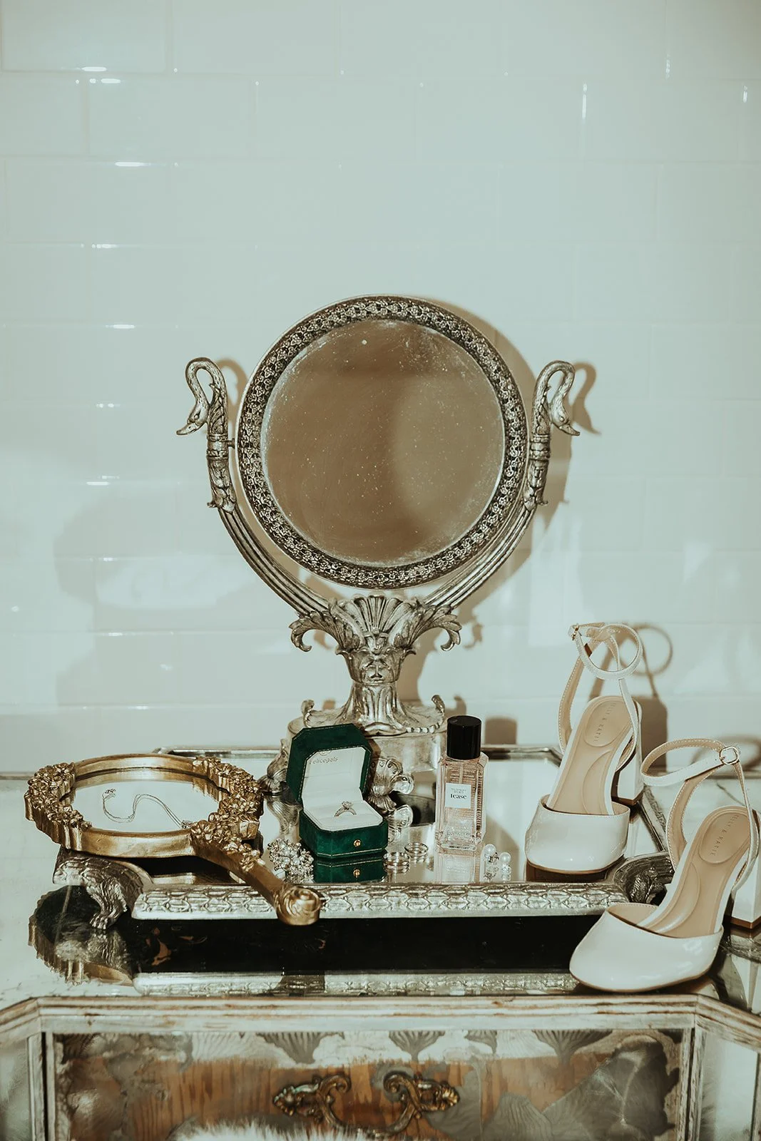 An ornate vintage vanity mirror with a decorative frame, surrounded by jewelry, a bottle of perfume, and high-heeled shoes on a decorative table against a white tiled wall.
