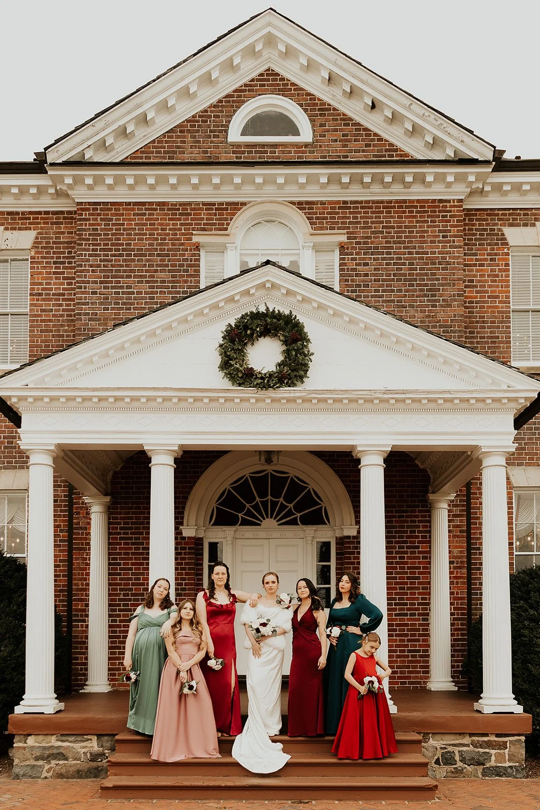 Group of women and a girl in bridesmaid and wedding dresses standing on the front porch of a brick house decorated with a Christmas wreath, posing for a photo.