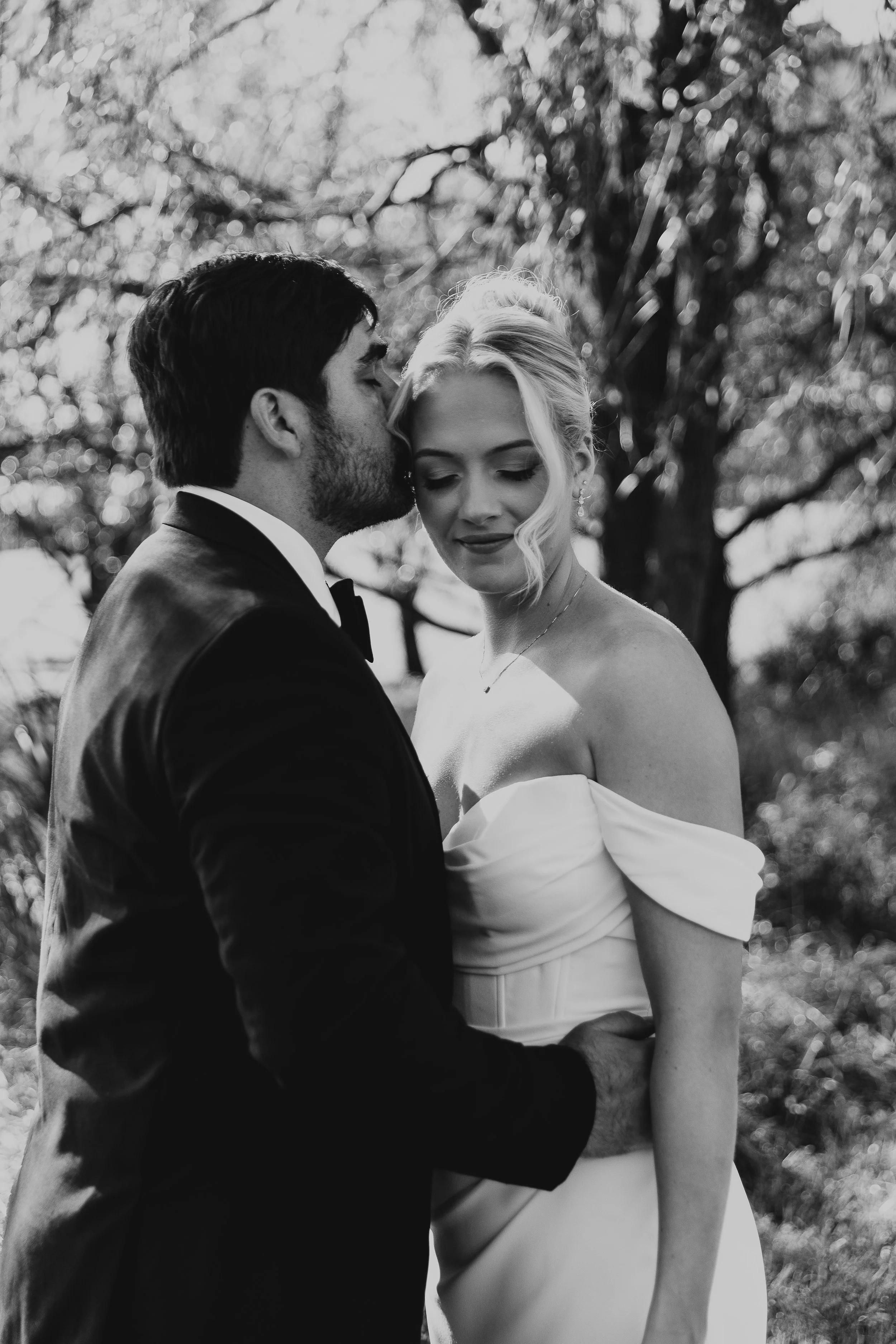 A black-and-white photo of a couple dressed in wedding attire, standing closely outdoors with trees in the background. The groom has dark hair and a beard, wearing a tuxedo with a bow tie, and is kissing the bride on her forehead. The bride has light