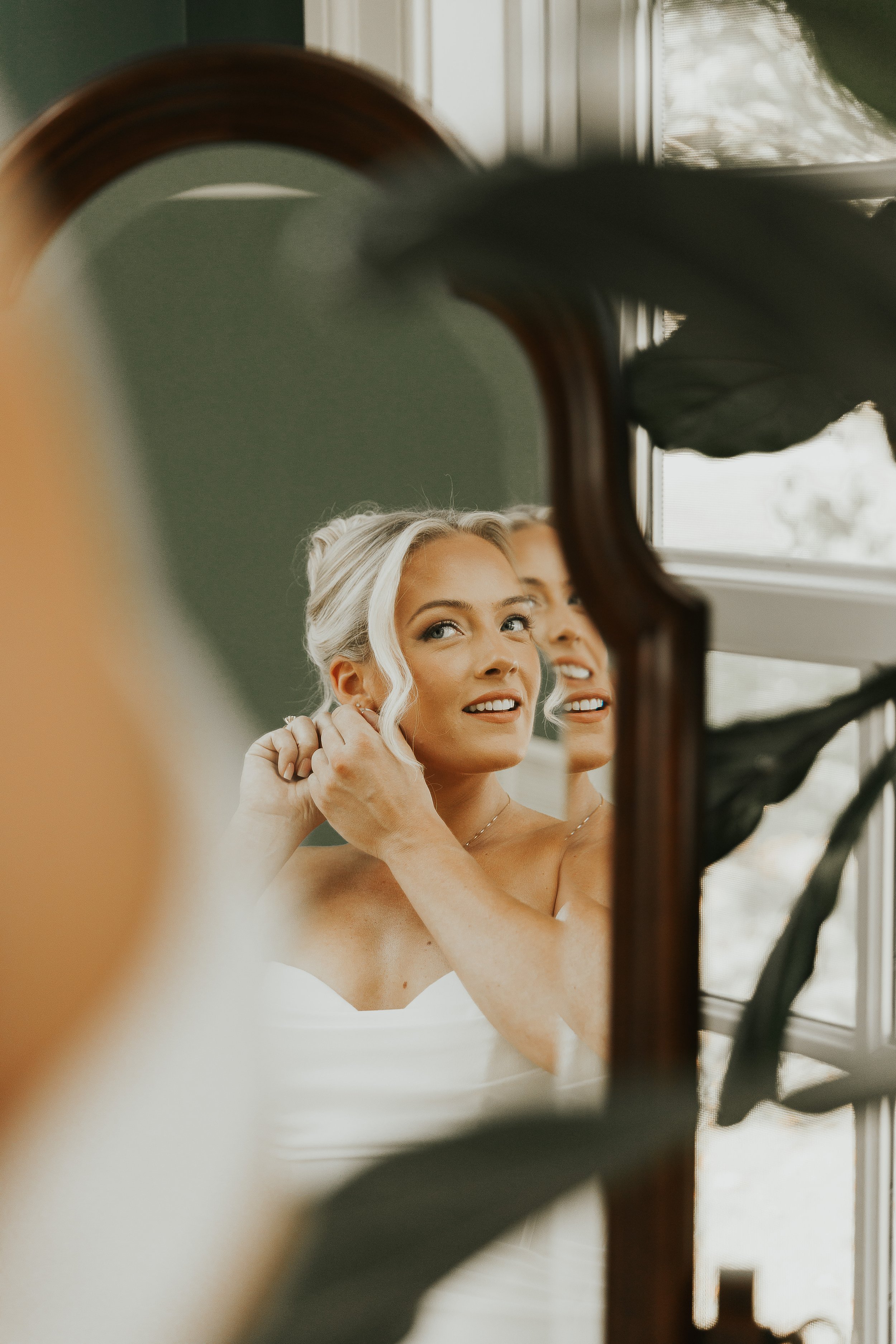 A young woman with platinum blonde hair styled in an updo is looking at herself in a mirror and smiling, as she adjusts her earrings. She is wearing a strapless white dress and is in a room with natural light coming through a window. The photo is tak